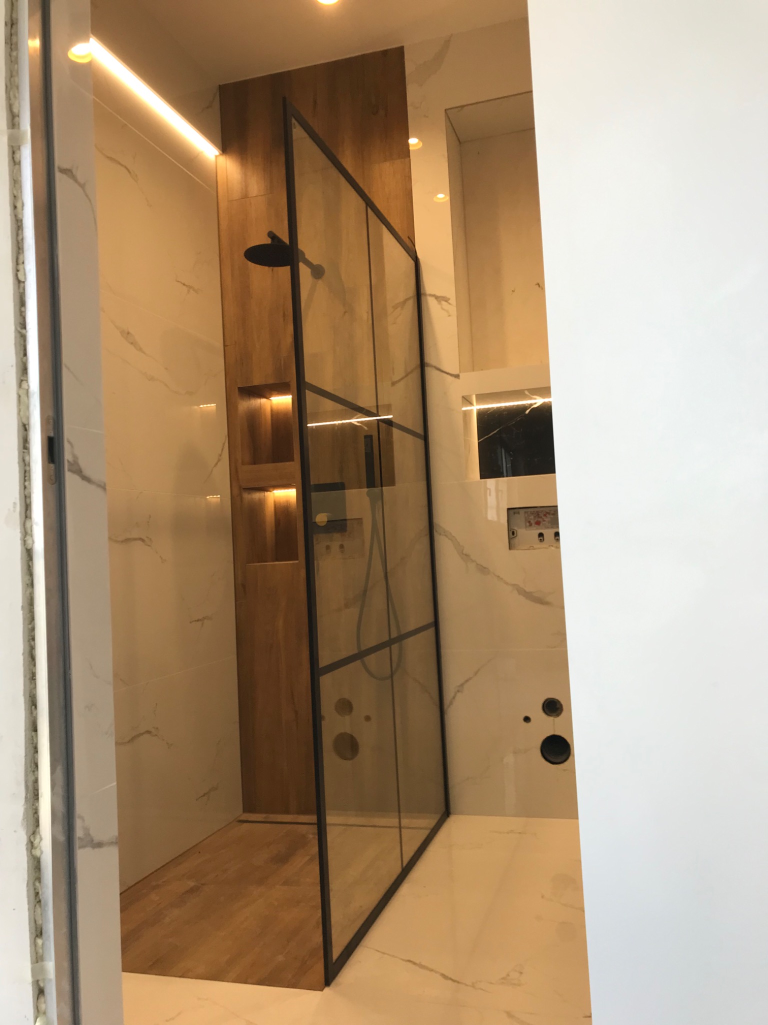 Nowoczesna kabina prysznicowa typu walk-in z czarnymi profilami, drewnianą ścianką z wnękami podświetlonymi ledami i marmurowymi płytkami w beżowych odcieniach.