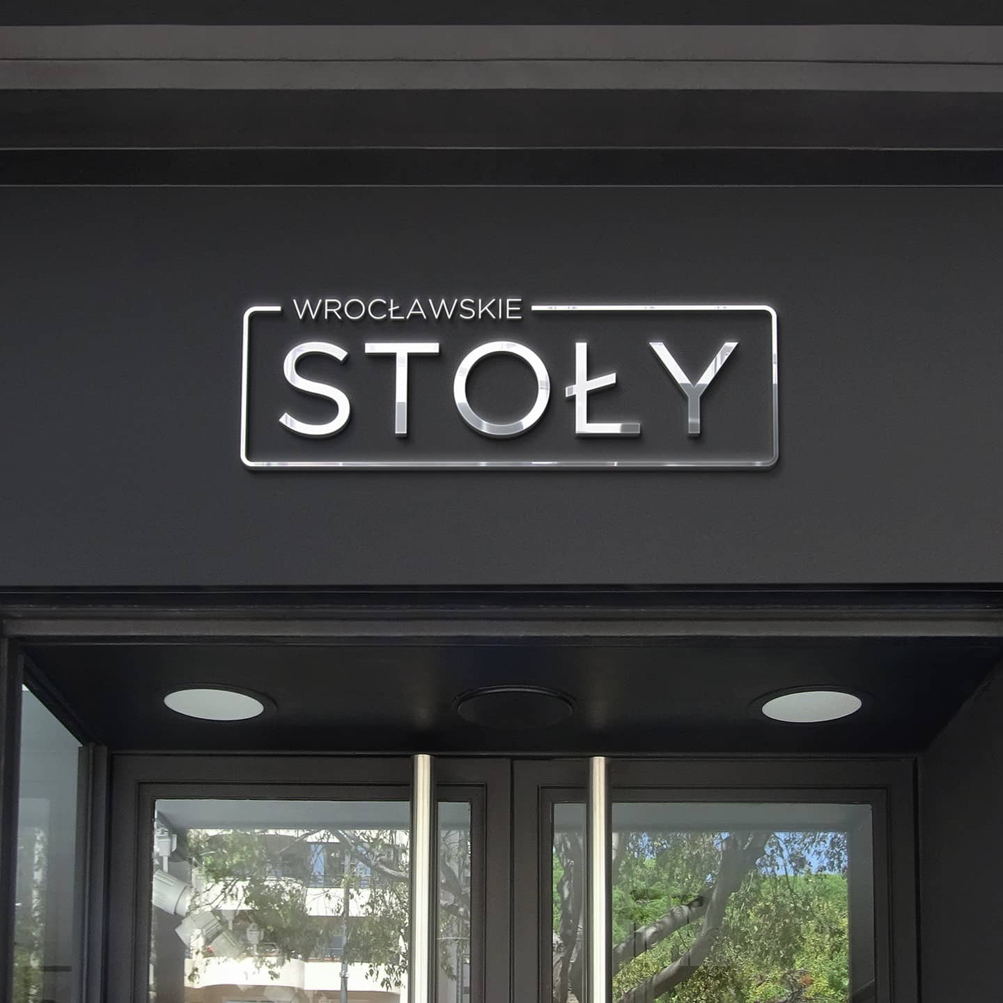 Elegancki szyld z nazwą 'Wrocławskie Stoły' w srebrnej ramce, zamontowany nad wejściem do budynku z ciemnymi drzwiami i oświetleniem punktowym.