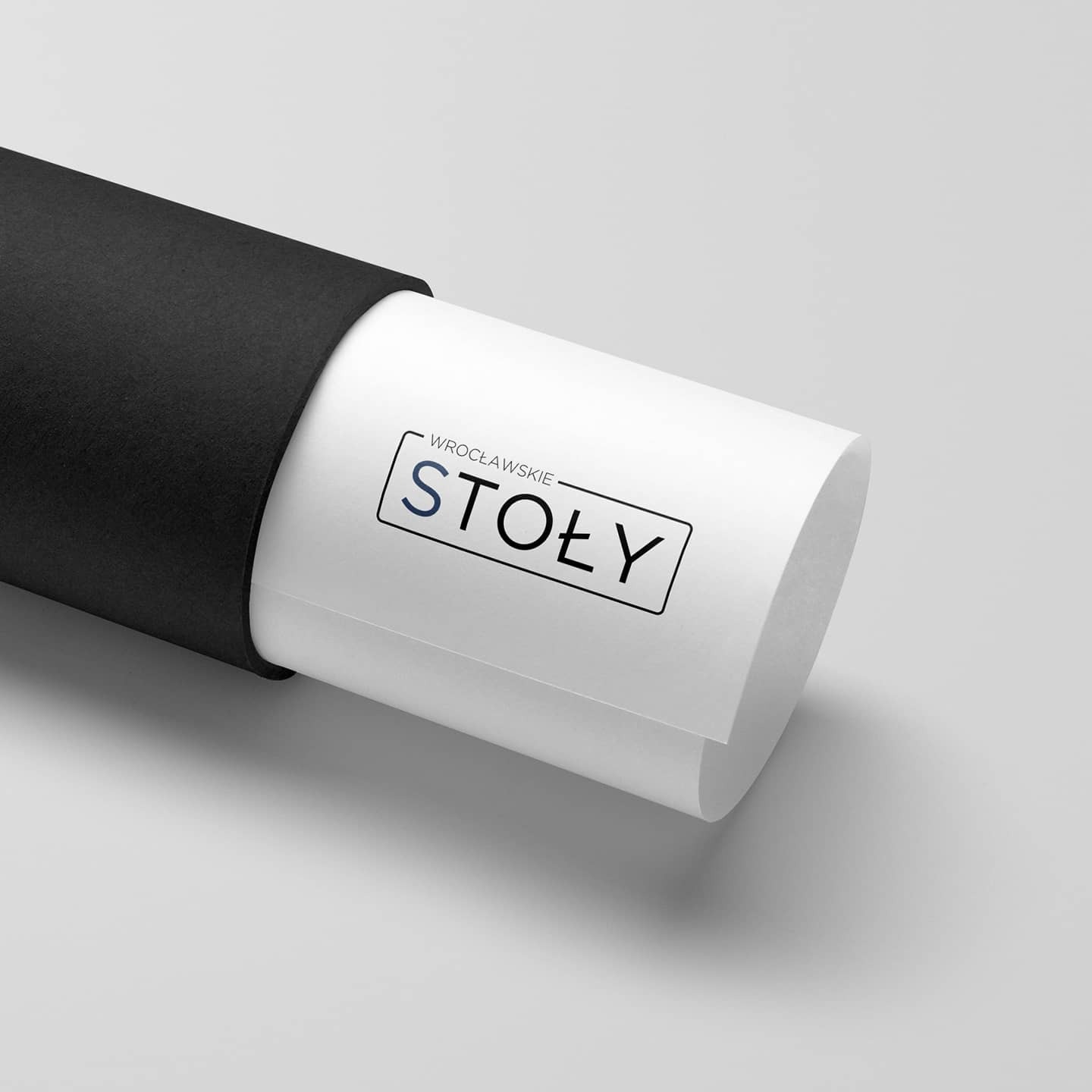 Grafika z napisem 'Wrocławskie Stoły' w czarnej ramce, wysuwająca się z czarnego etui na jasnoszarym tle.
