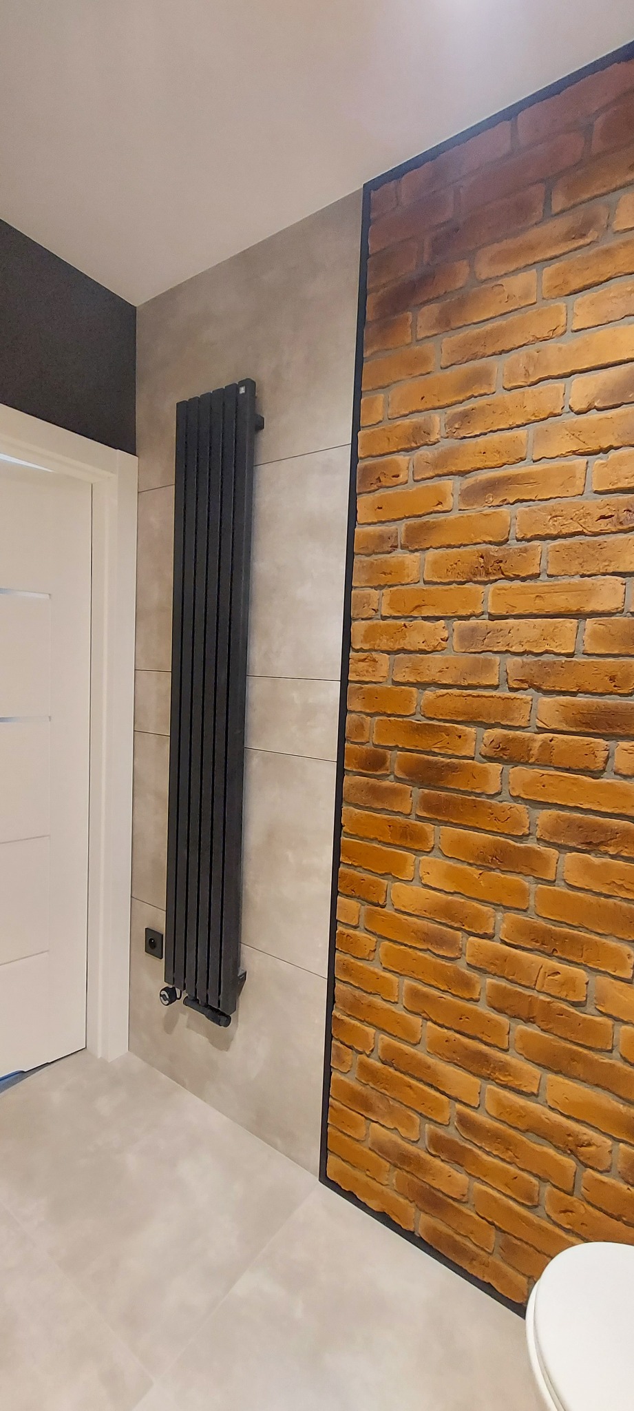 Wnętrze łazienki z grzejnikiem panelowym na ścianie z płytek imitujących beton, obok ściana z cegły klinkierowej w odcieniach brązu i pomarańczy, widoczny fragment białej miski WC.
