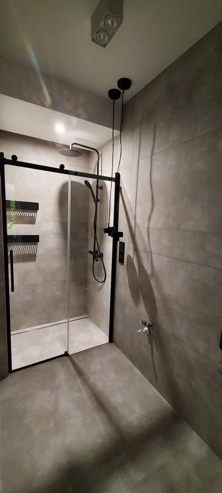 Nowoczesna kabina prysznicowa z czarnymi ramami i deszczownicą, szare płytki na ścianach i podłodze, widoczny fragment instalacji oświetleniowej na suficie.