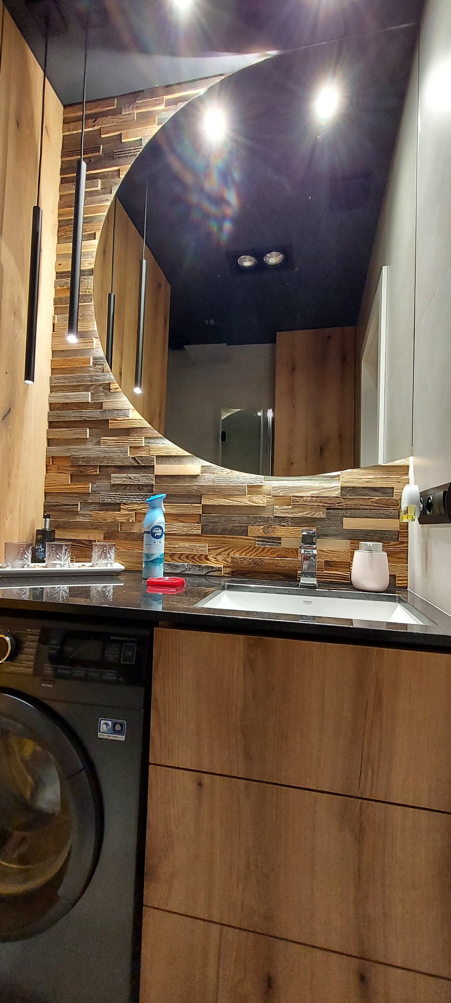 Nowoczesna łazienka z umywalką na blacie z ciemnego kamienia, obok pralka, na ścianie lustro z podświetleniem i dekoracyjna okładzina z drewnianych elementów różnej wielkości, wiszące lampy