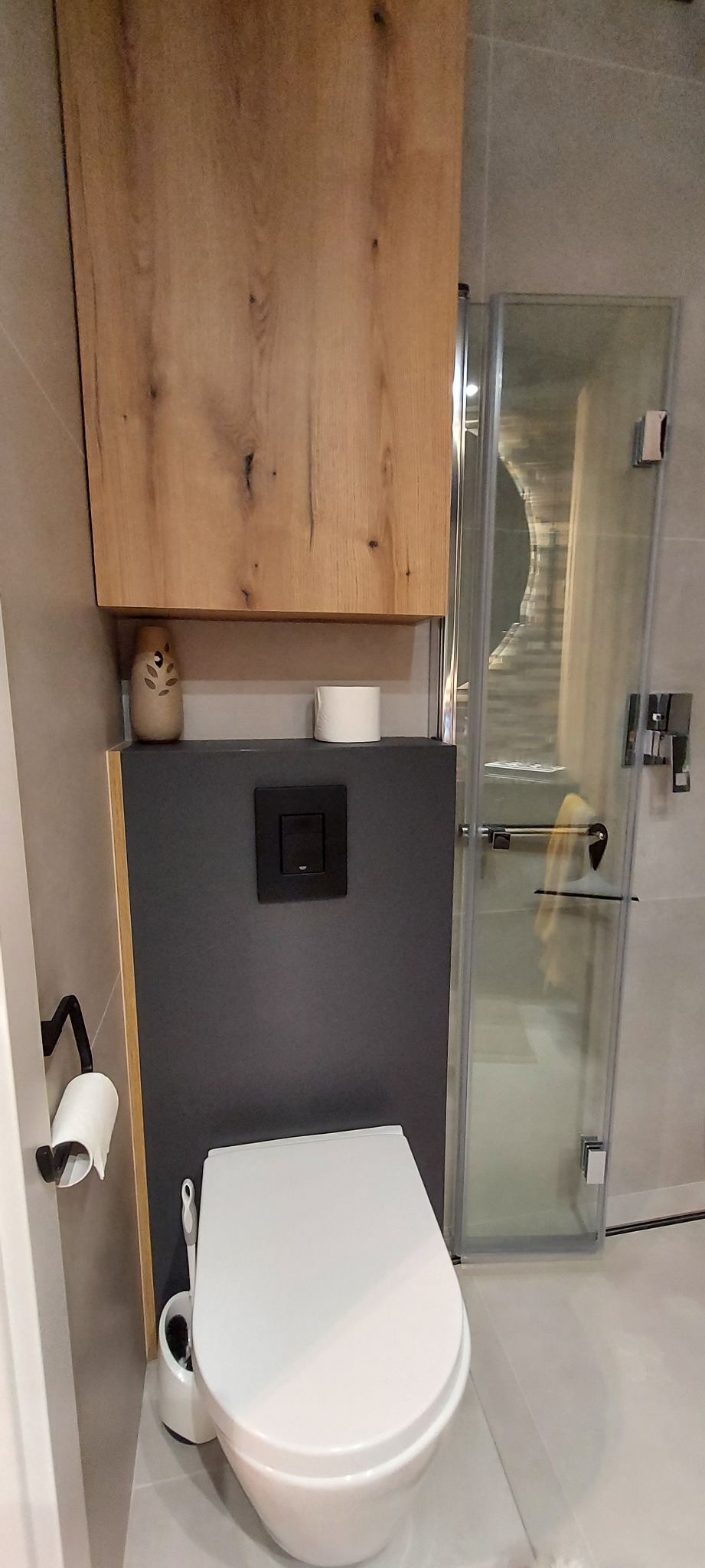 Nowoczesna toaleta podwieszana z szarą ścianką, drewnianą szafką nad nią, obok kabina prysznicowa ze srebrnymi okuciami i widocznym ręcznikiem wewnątrz.