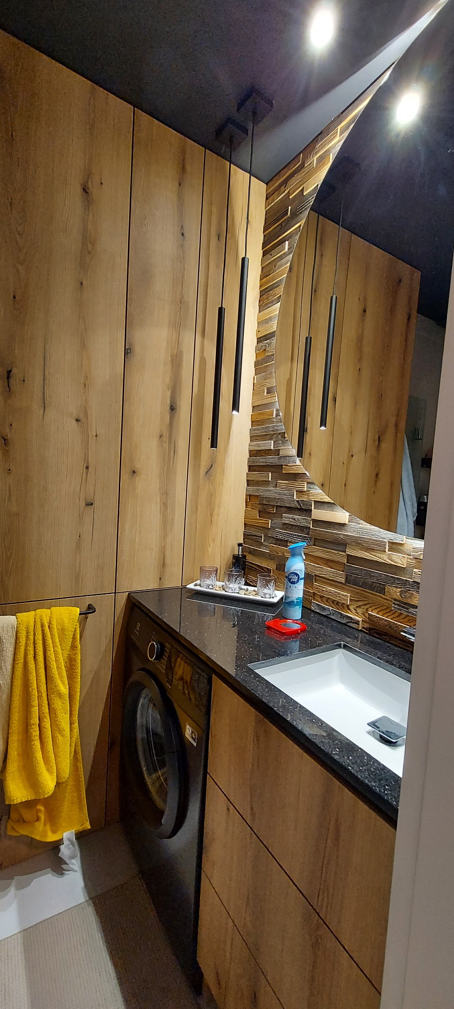 Nowoczesna łazienka z drewnianą zabudową, czarnym blatem, umywalką, pralką, okrągłym lustrem z drewnianą ramą i wiszącymi lampami.