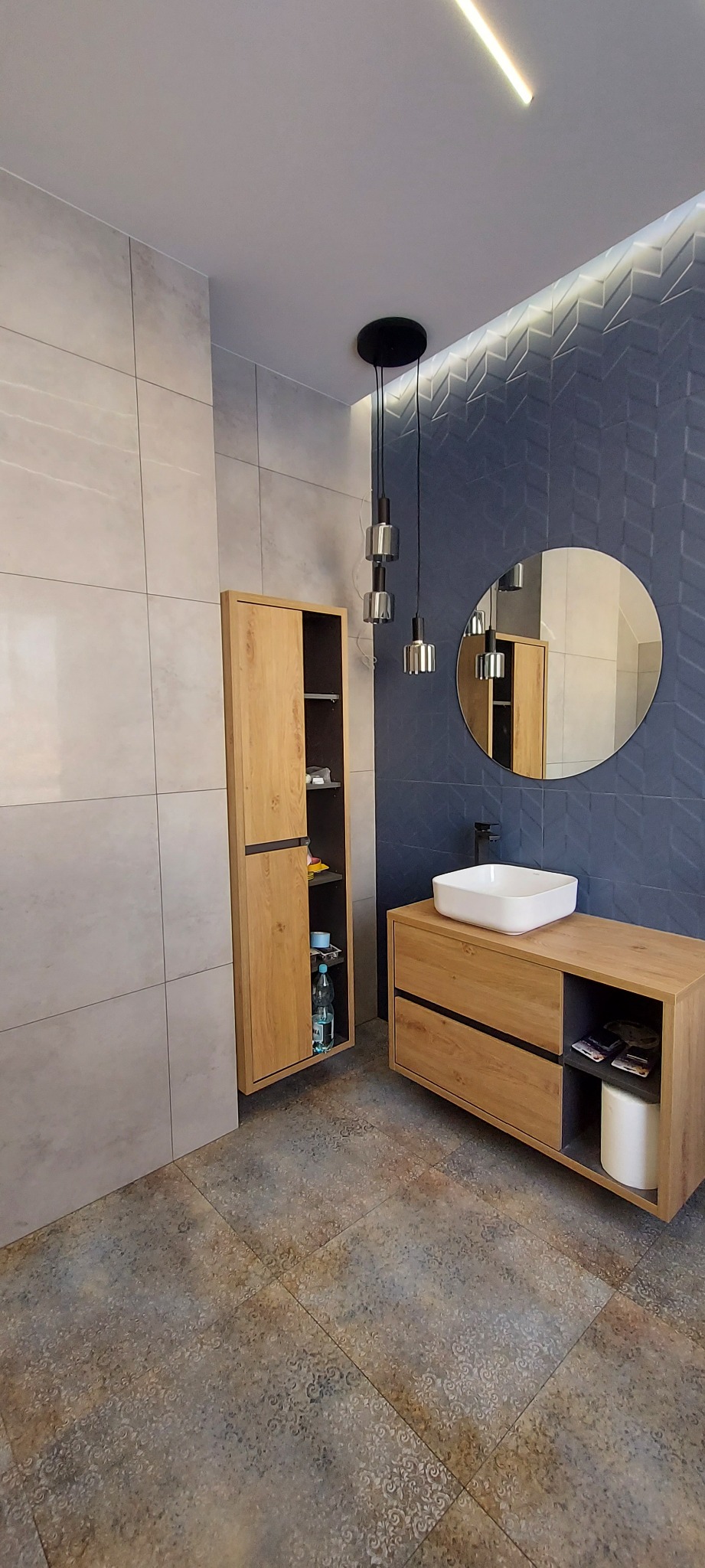 Nowoczesna łazienka z drewnianą szafką pod umywalką, lustrem i szafką wiszącą z częściowo otwartymi drzwiczkami, ukazującymi butelkę wody w środku, na tle niebieskiej ściany z wzorem jodełki...