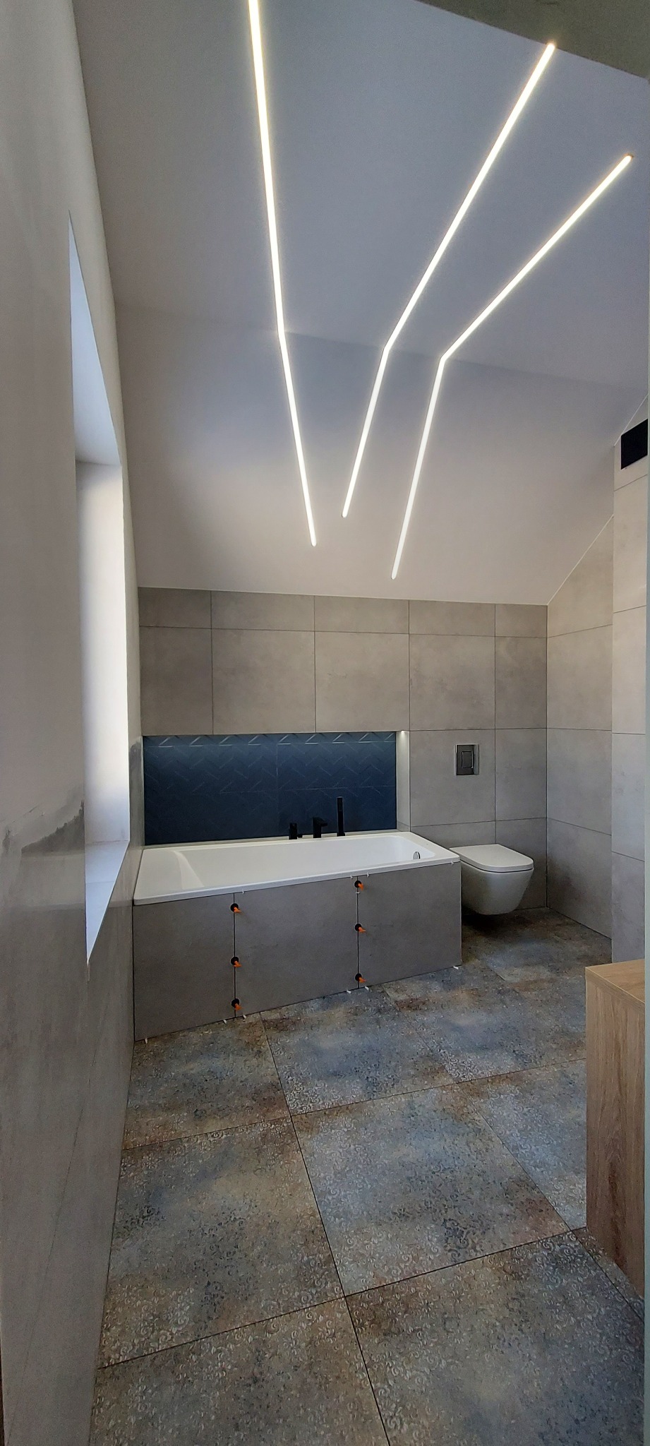 Nowoczesna łazienka z podświetlanym sufitem, szarymi płytkami i dekoracyjną ścianką nad wanną, widoczne krzyżyki dystansowe na obudowie wanny.