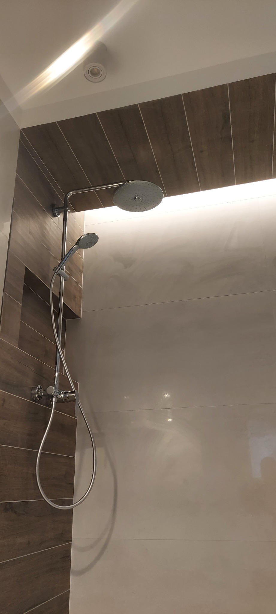 Nowoczesna kabina prysznicowa z deszczownicą i dodatkową słuchawką, wykończona płytkami imitującymi drewno na jednej ścianie i jasnymi, gładkimi płytkami na pozostałych ścianach, oświetlona ledowym...