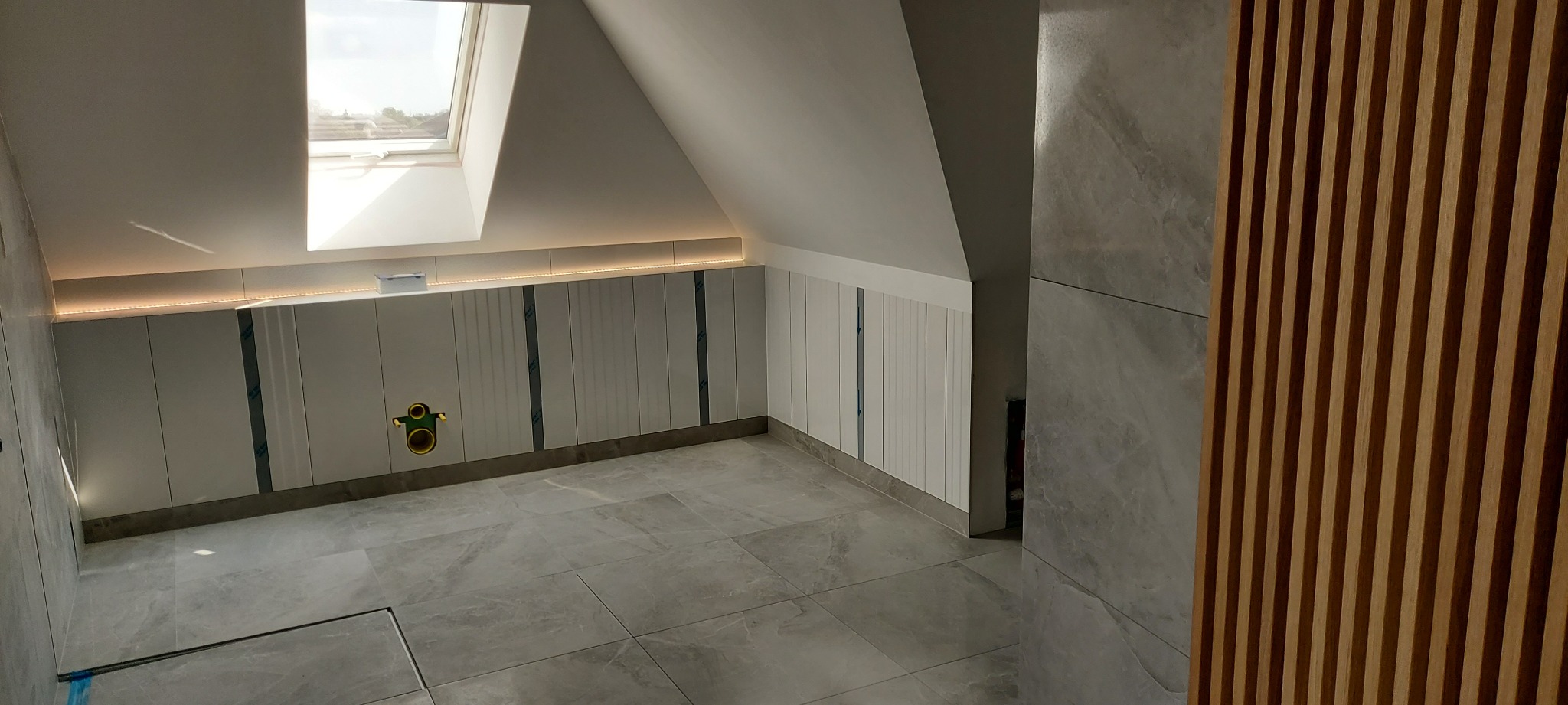 Nowoczesna łazienka na poddaszu z szarymi płytkami podłogowymi i ściennymi imitującymi kamień, białe szafki pod skosem ze zintegrowanym oświetleniem LED oraz drewniane panele dekoracyjne.