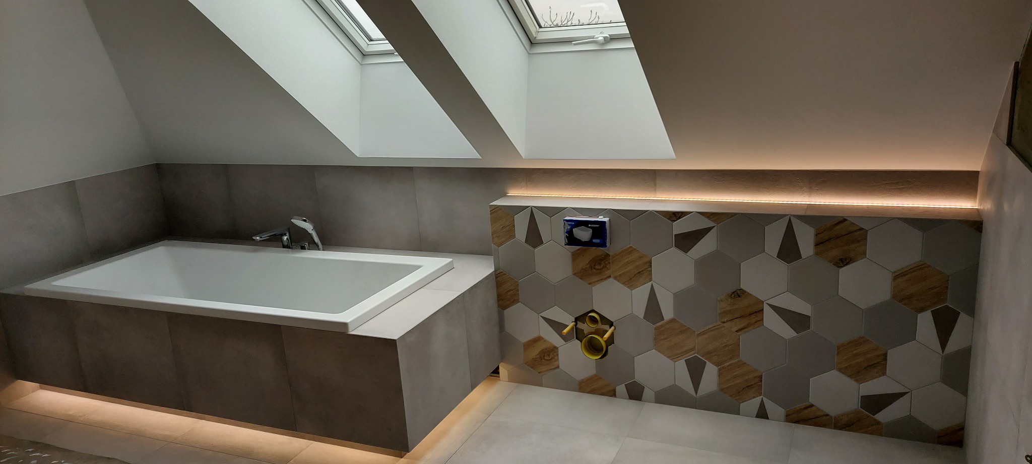 Nowoczesna łazienka na poddaszu z oknem dachowym, białą wanną obudowaną szarą płytką, heksagonalnymi płytkami na ścianie z drewnopodobnym wzorem i podświetleniem LED.