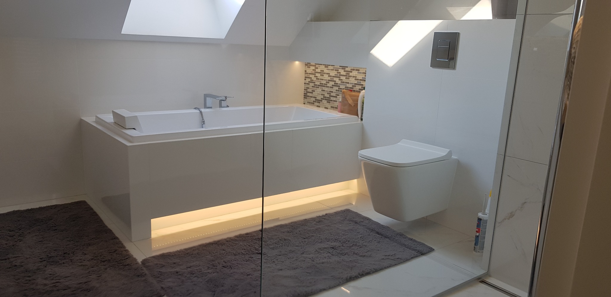 Nowoczesna, jasna łazienka z podświetlaną wanną, wiszącą toaletą i szklaną kabiną prysznicową, widok zza szyby, dywaniki łazienkowe w odcieniach szarości.