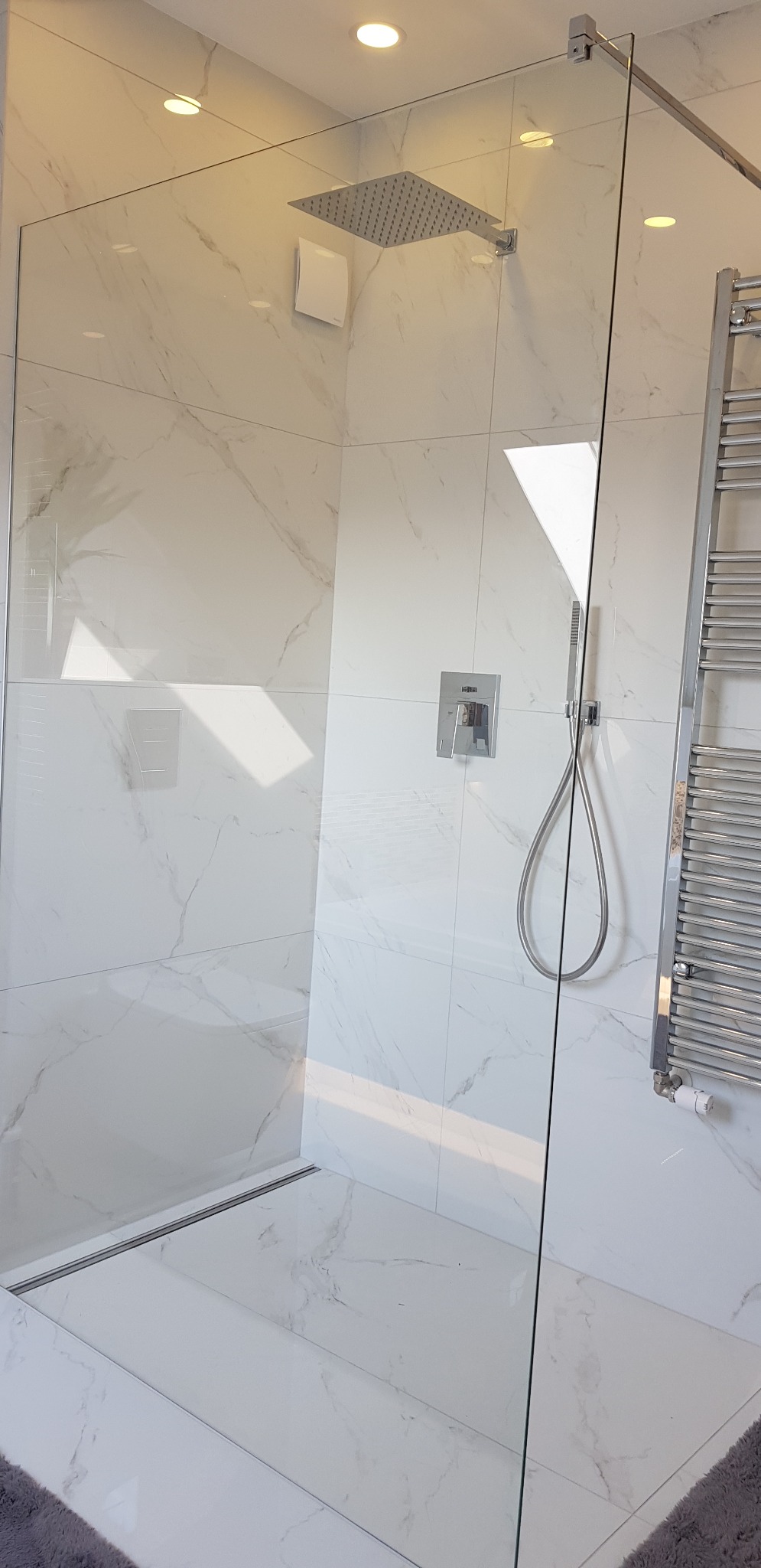 Nowoczesna kabina prysznicowa typu walk-in z deszczownicą i grzejnikiem drabinkowym, wykończona białymi płytkami imitującymi marmur.