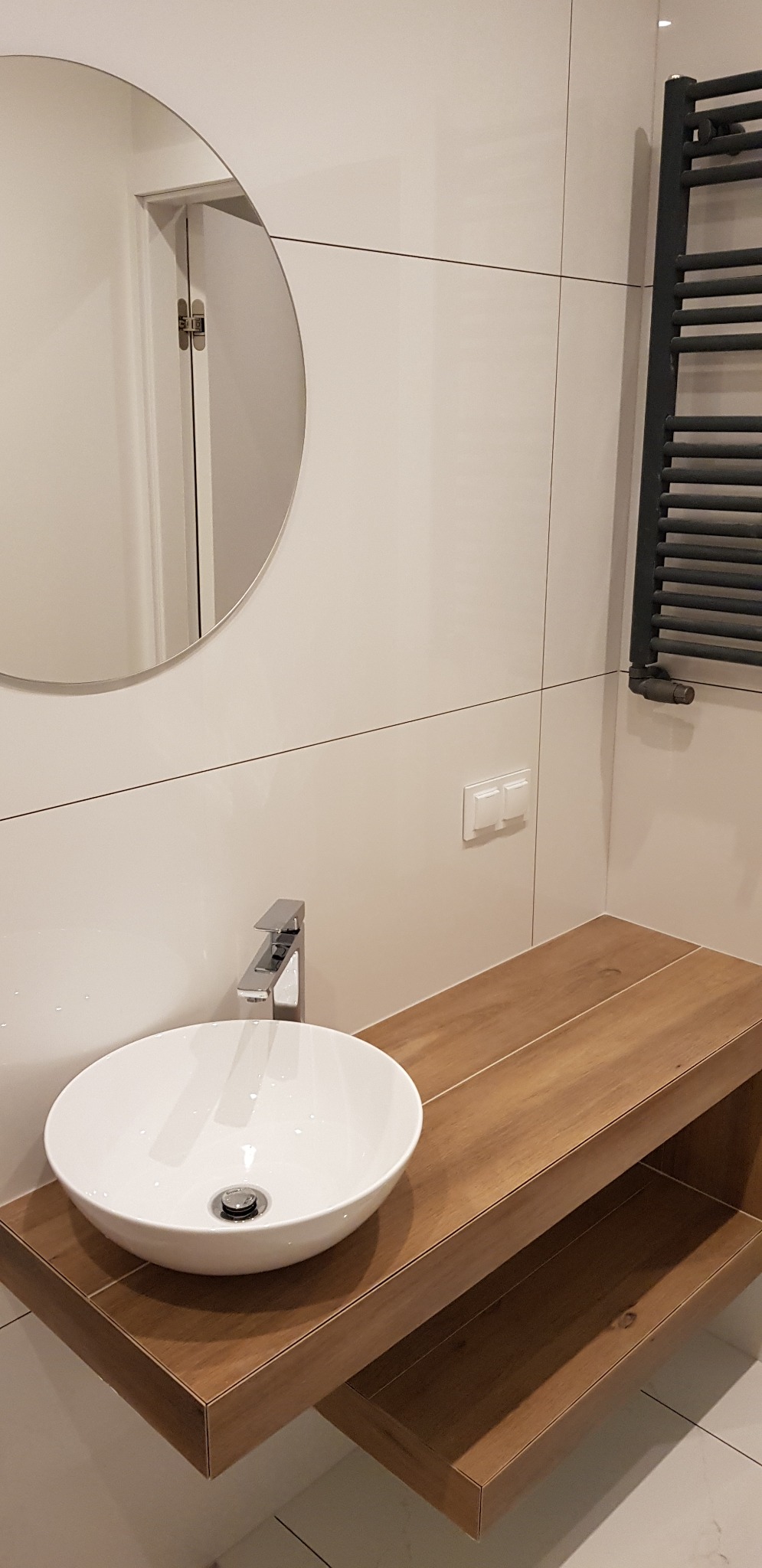 Nowoczesna łazienka z okrągłym lustrem, białą umywalką nablatową i drewnopodobnym blatem z półką, wykończona białymi płytkami.