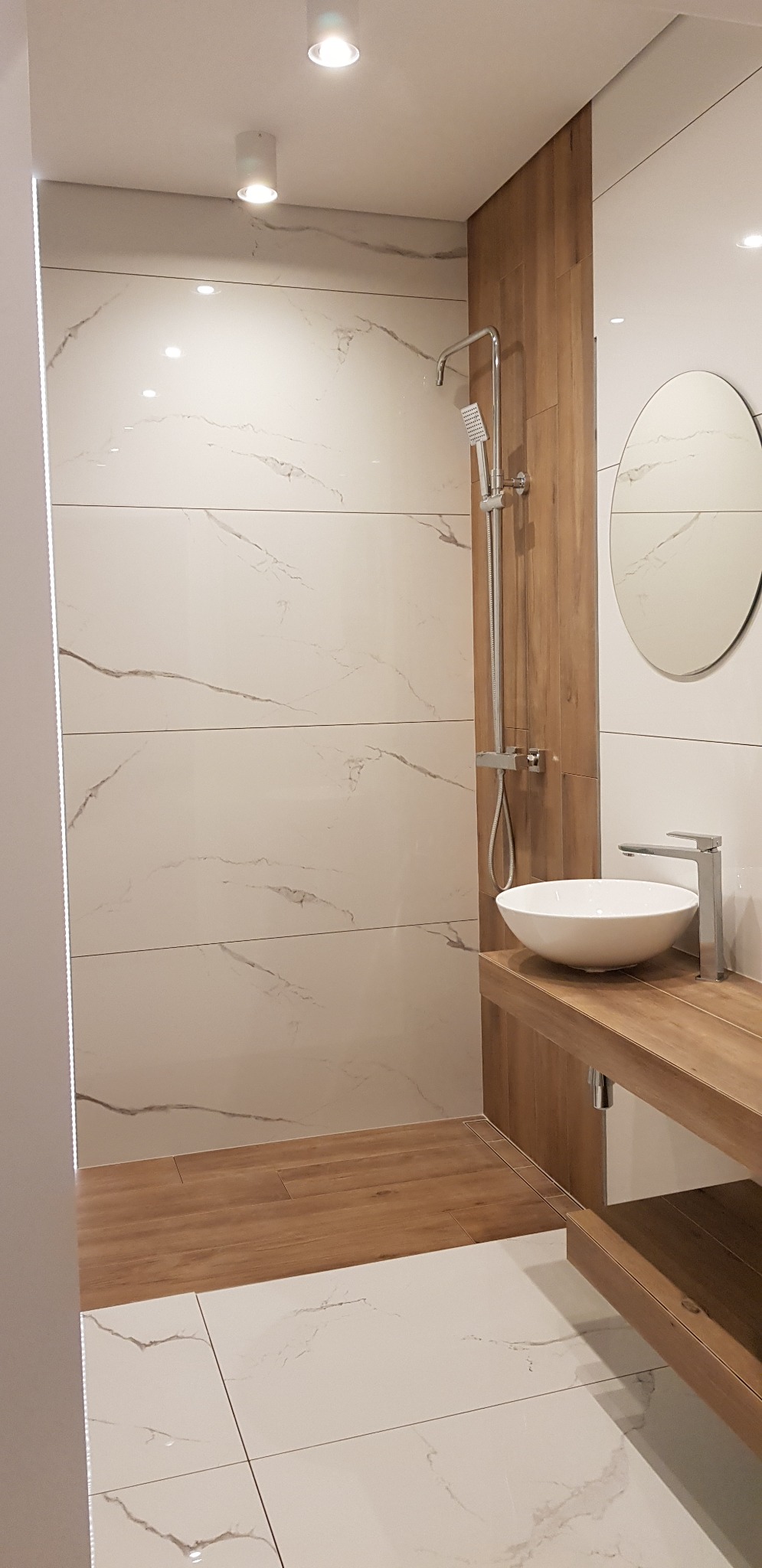 Nowoczesna łazienka z kabiną prysznicową typu walk-in, umywalką nablatową na drewnianym blacie, owalnym lustrem i białymi płytkami imitującymi marmur.