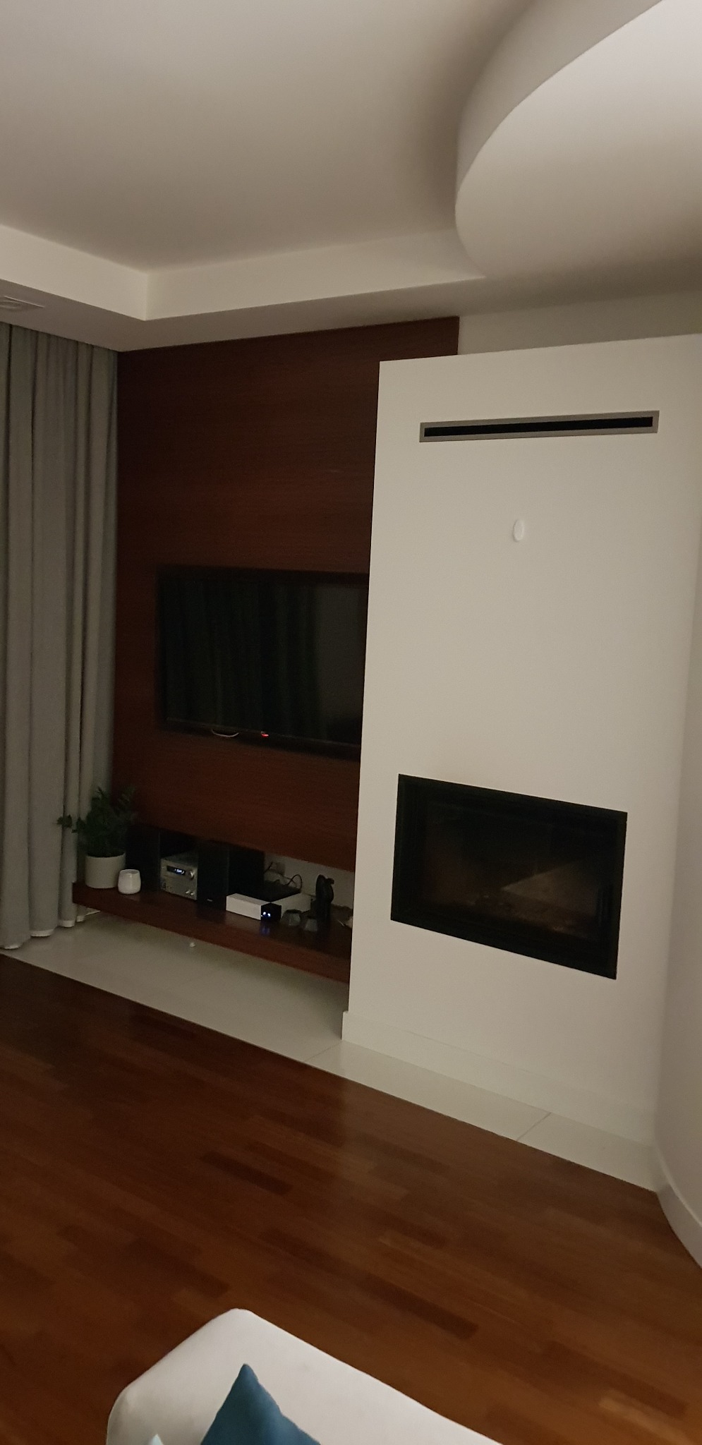 Nowoczesny salon z drewnianą ścianą, telewizorem i białym kominkiem z czarnym wkładem, minimalistyczny design, oświetlenie sufitowe.