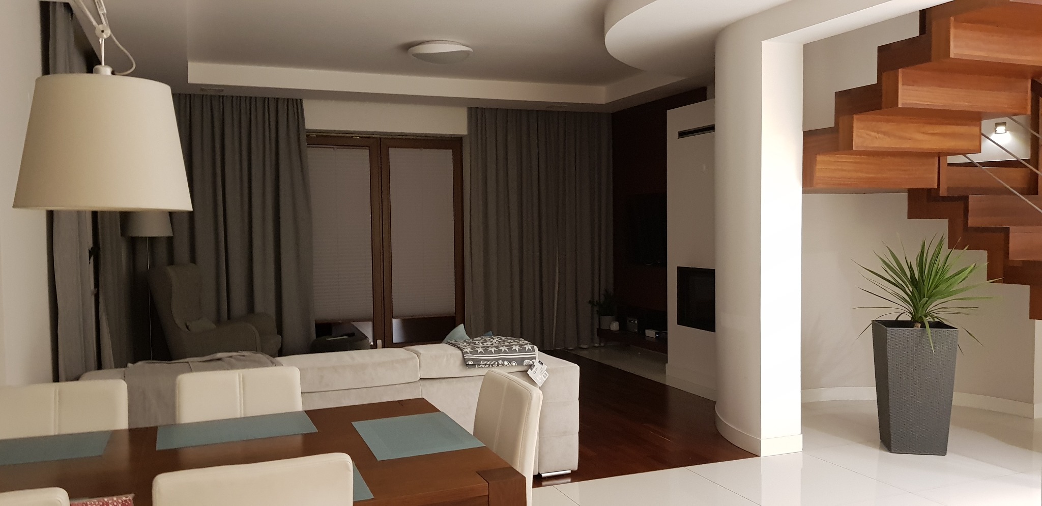 Jasne wnętrze domu z drewnianymi schodami spiralnymi, widok na salon z beżową sofą i szarymi zasłonami, oraz jadalnię z drewnianym stołem i białymi krzesłami, podłoga z białych płytek.