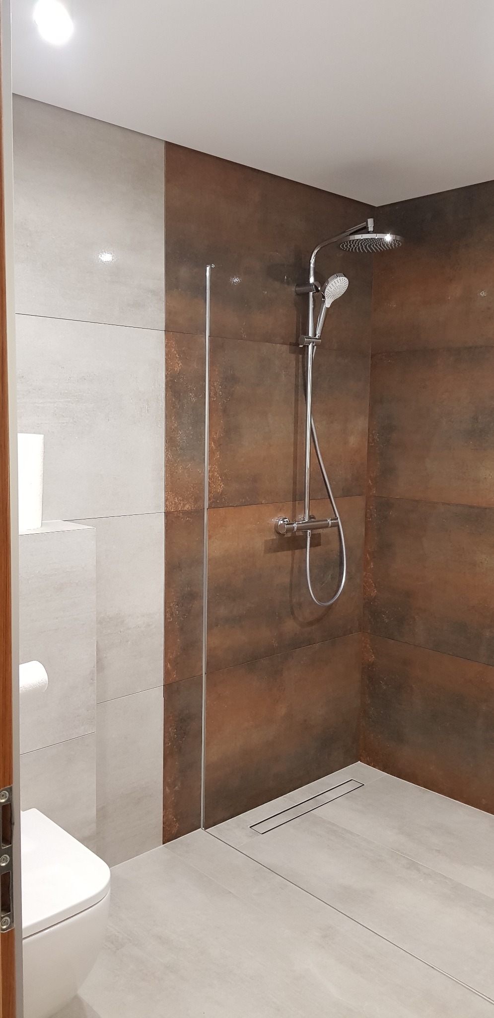 Nowoczesna kabina prysznicowa z deszczownicą, wykończona płytkami w odcieniach szarości i rdzy, widoczny odpływ liniowy w podłodze i toaleta w tle.