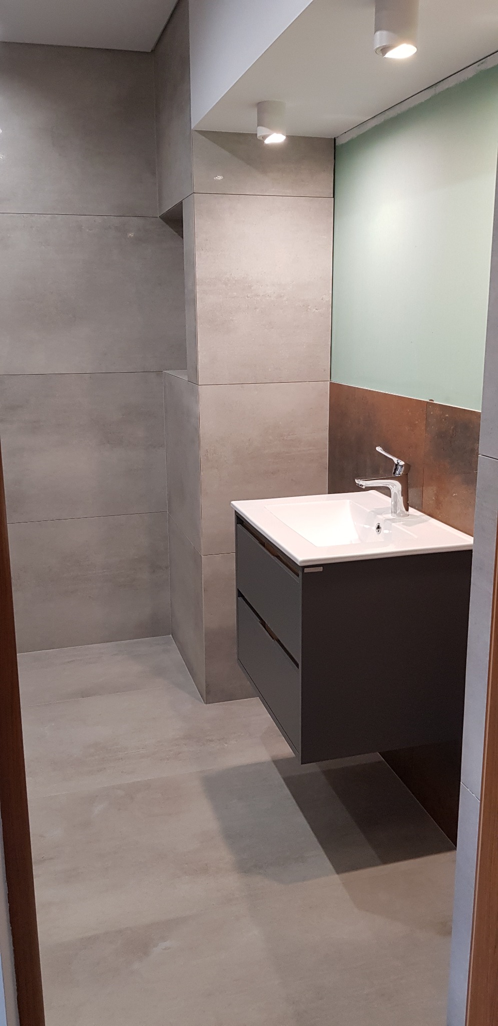 Nowoczesna łazienka z szarymi płytkami imitującymi beton, wisząca szafka umywalkowa z ciemnoszarymi frontami i białą umywalką oraz chromowaną baterią.