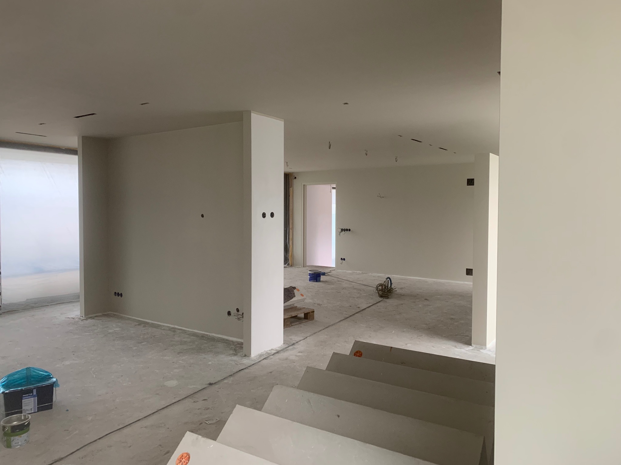 Wnętrze w trakcie prac wykończeniowych: gładkie, pomalowane na jasny kolor ściany, surowy beton na podłodze, widoczne puszki elektryczne, narzędzia malarskie i paleta w tle.