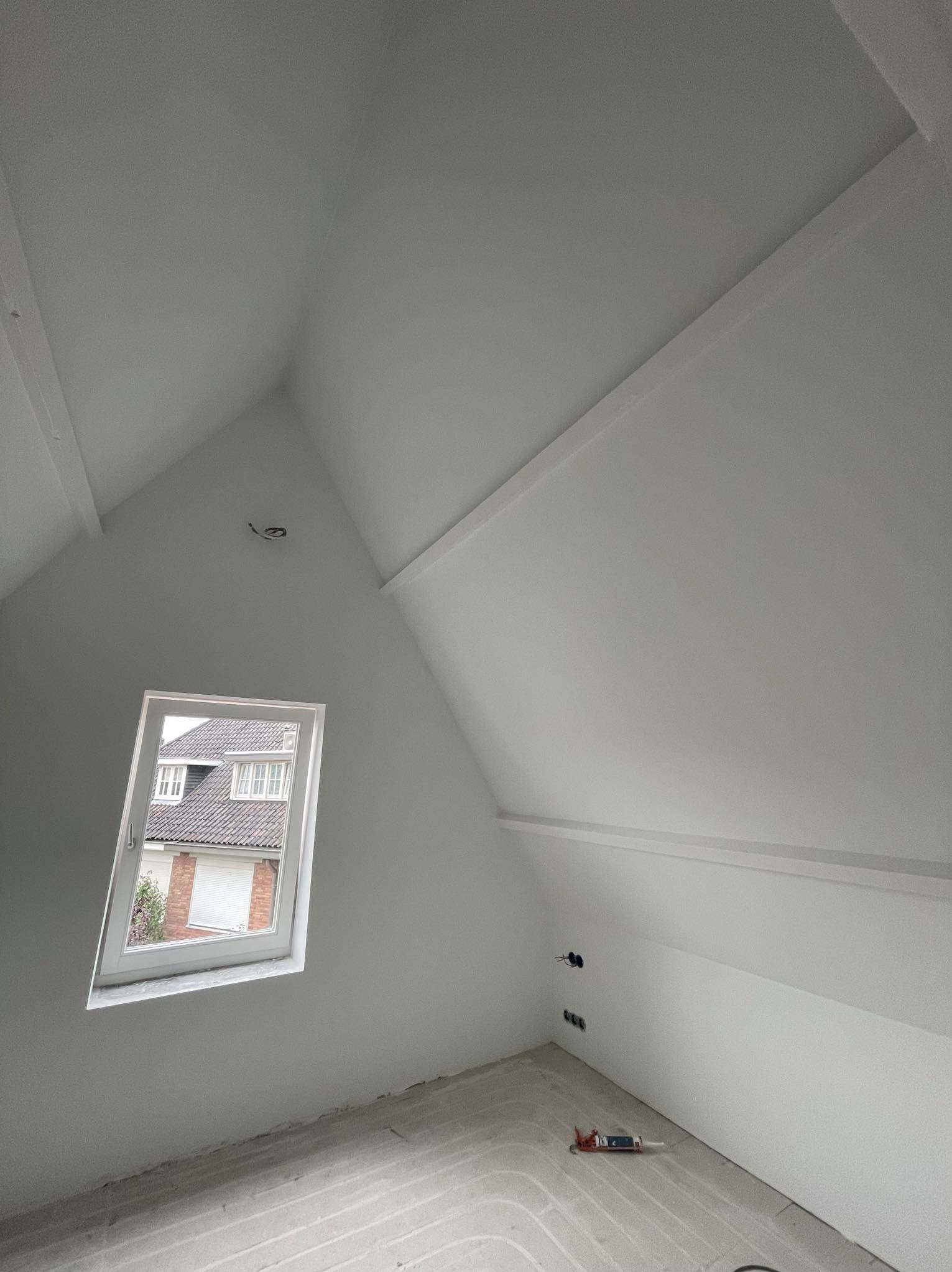 Poddasze po pracach wykończeniowych, świeżo pomalowane na biało, z widocznymi elementami instalacji elektrycznej i oknem dachowym z widokiem na sąsiedni dom.