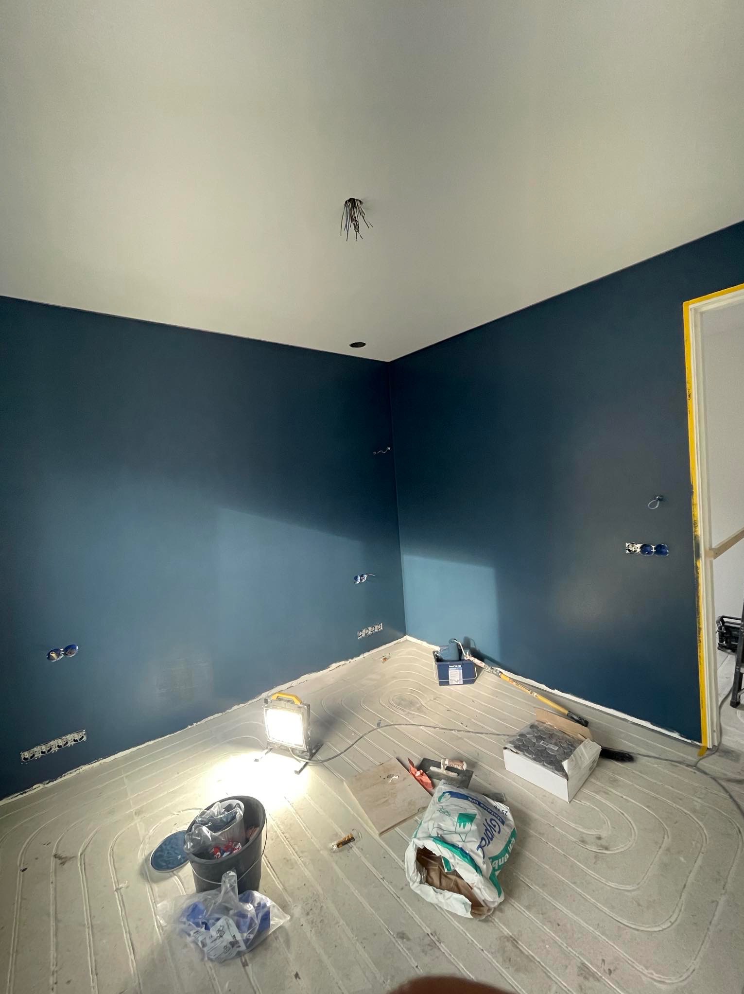 Pokój po świeżym malowaniu na ciemny odcień niebieskiego, z widocznymi elementami instalacji elektrycznej, podłogą z ogrzewaniem podłogowym i narzędziami malarskimi w trakcie prac wykończeniowych.
