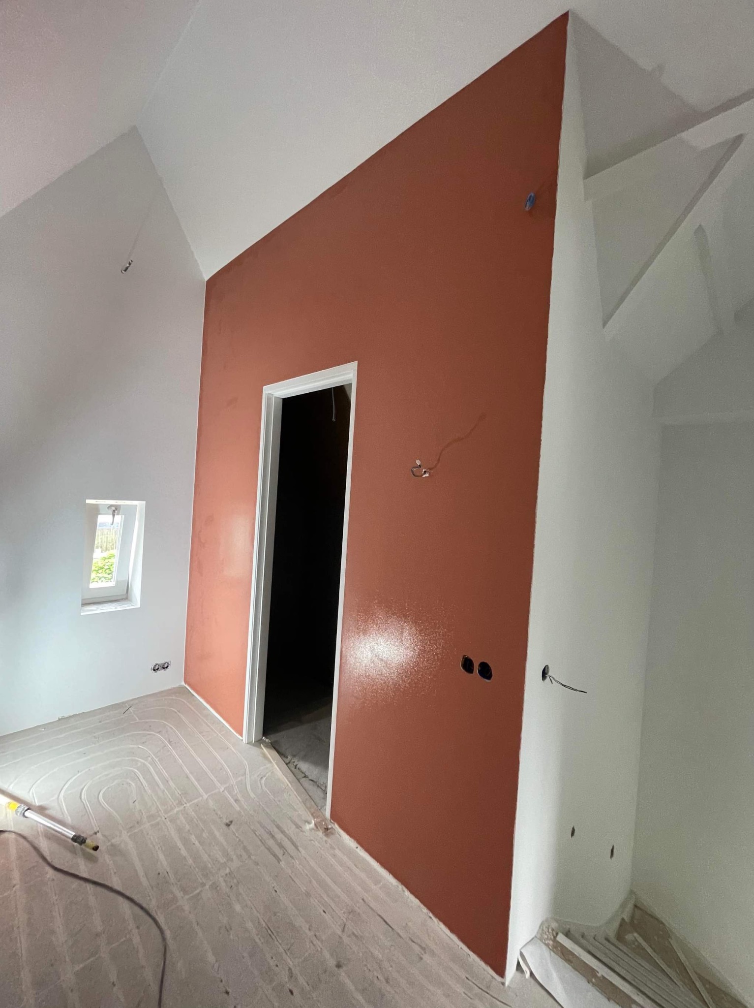 Pokój w trakcie prac wykończeniowych: jedna ściana pomalowana na terakotowy kolor, pozostałe białe, widoczne instalacje elektryczne, ogrzewanie podłogowe w wylewce.