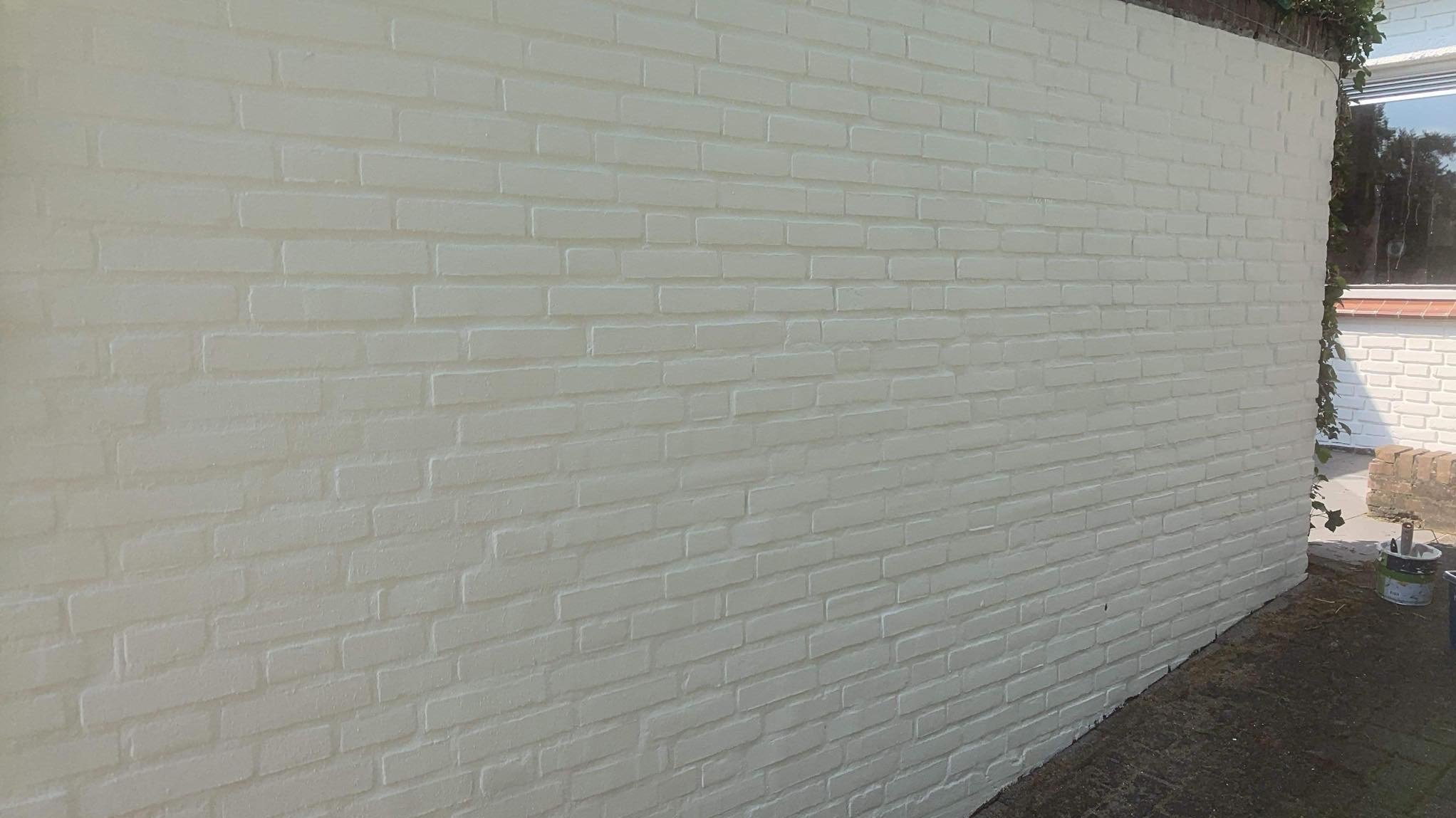 Świeżo pomalowany na kremowo ceglany mur zewnętrzny, z widocznym wiadrem z farbą w tle i fragmentem okna z zielenią.