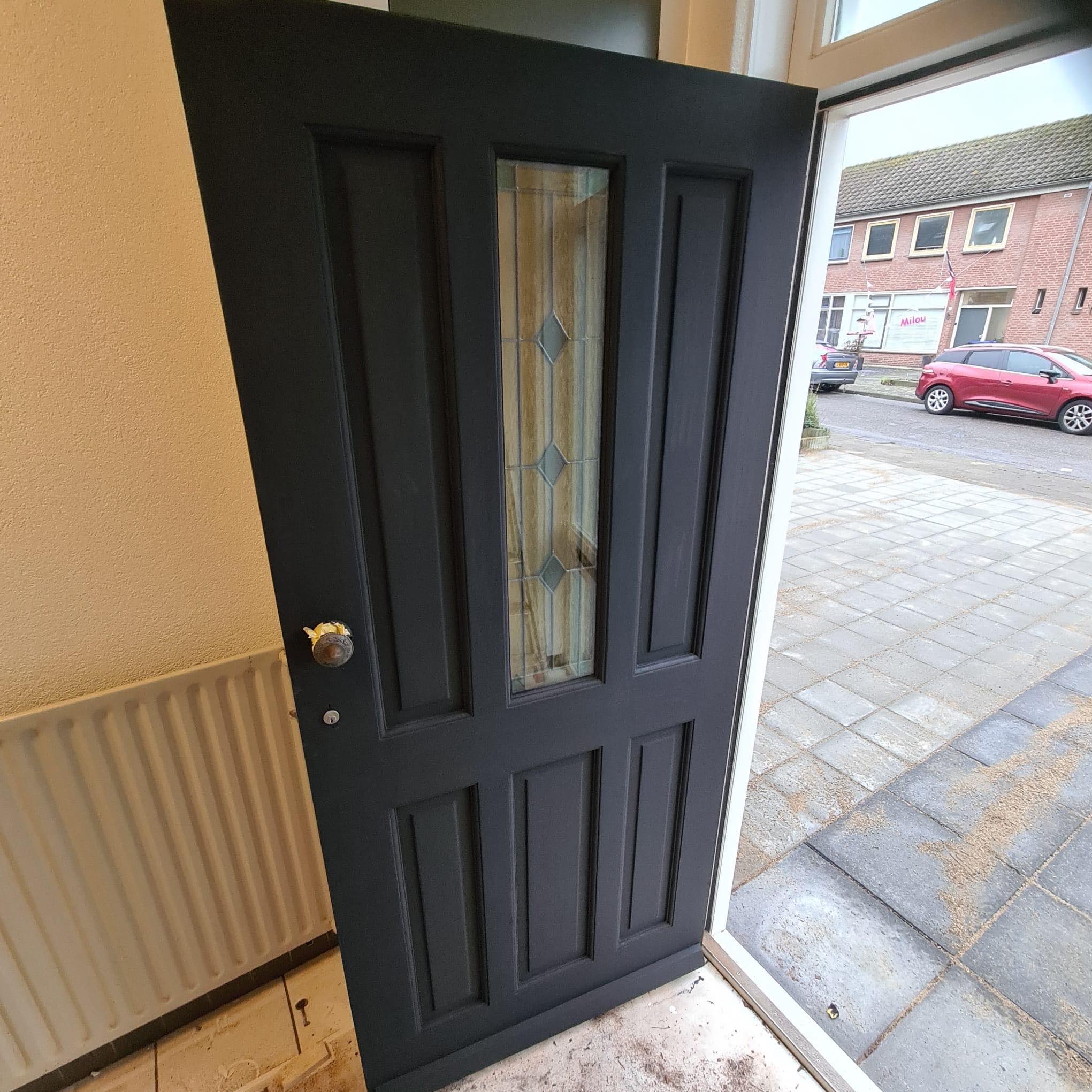 Ciemnoszare drzwi wejściowe z szybą z dekoracyjnym szkłem, widok z wnętrza na kostkę brukową i budynki mieszkalne