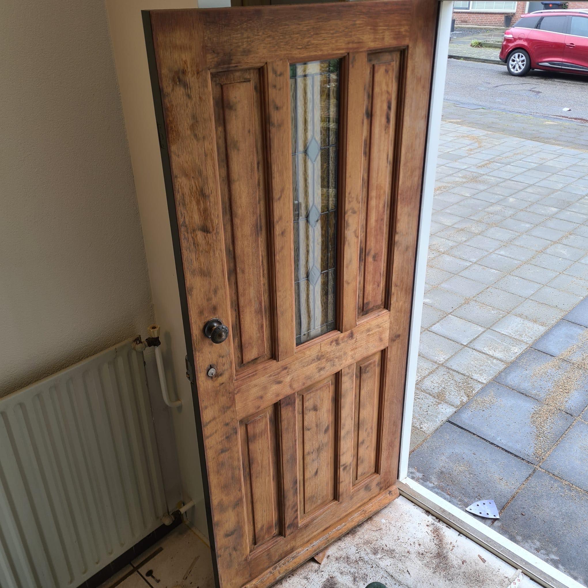 Drewniane drzwi wejściowe z szybą, częściowo odnowione, z widocznymi śladami starej farby, w trakcie prac renowacyjnych, z widocznym grzejnikiem i fragmentem podłogi.