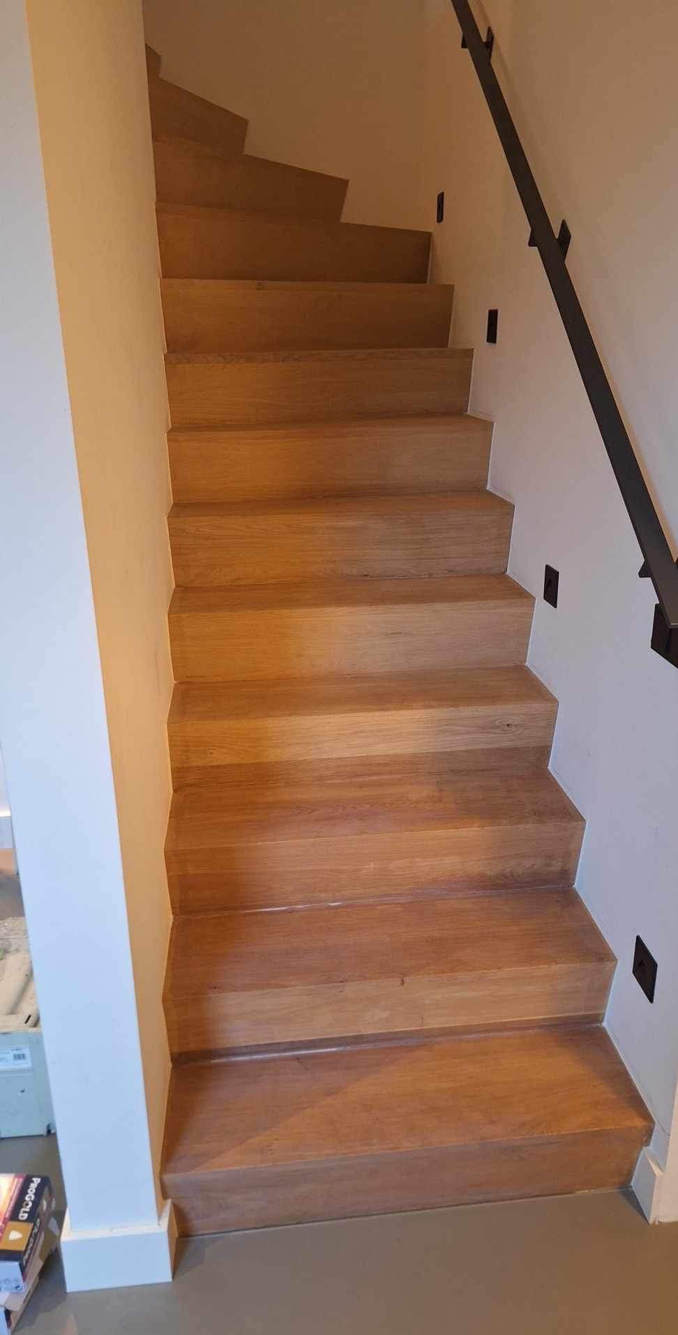 Schody drewniane o jasnym odcieniu, z czarną metalową balustradą i kontaktami przy schodach na białej ścianie.