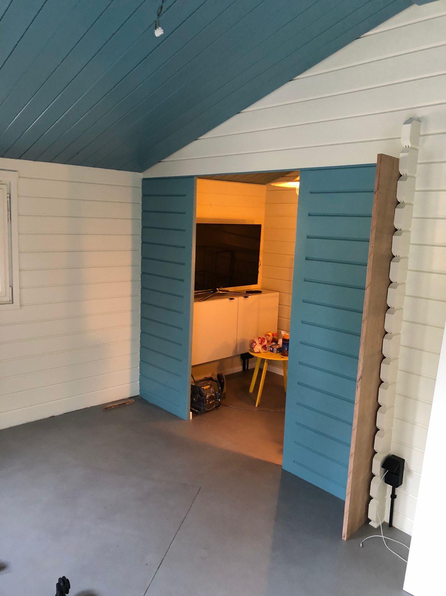 Pomalowane na biało deski boazerii ściennej, szary betonowy podkład, błękitny sufit z desek i przesuwne drzwi w kolorze błękitu prowadzące do pokoju z telewizorem i żółtym stolikiem.