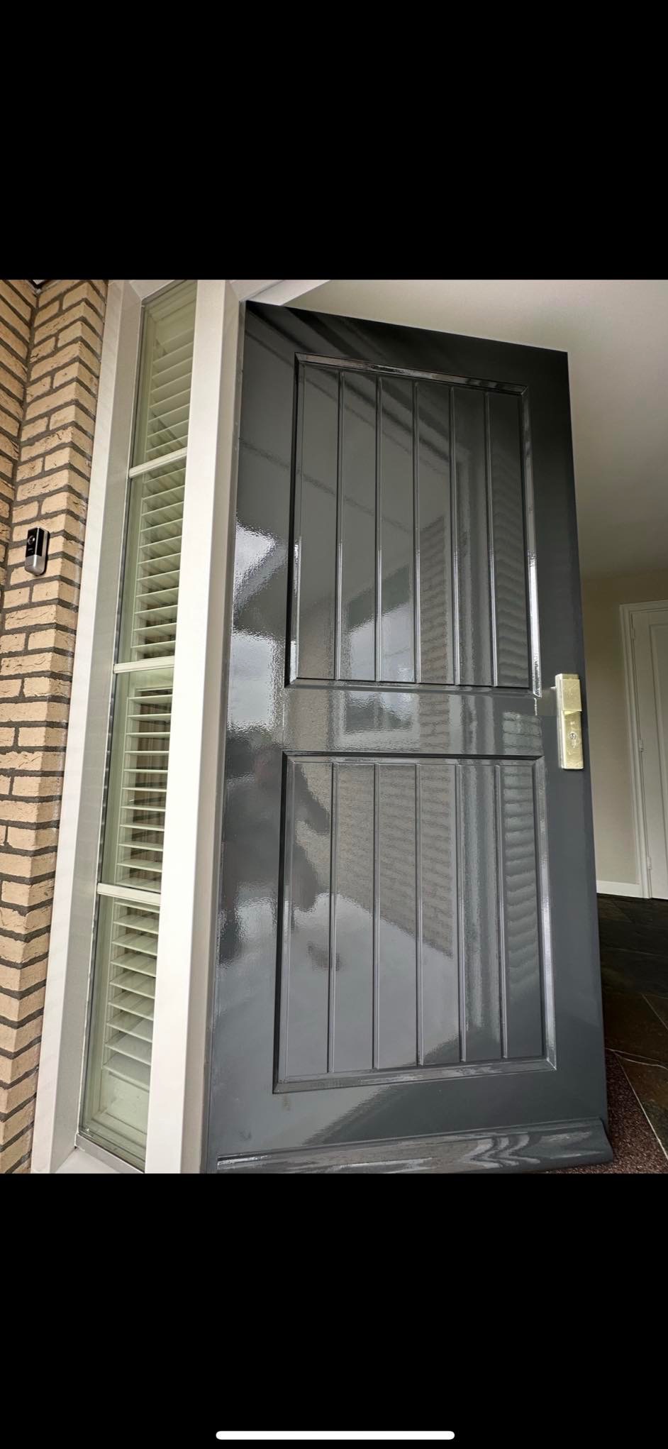 Świeżo pomalowane na szaro drzwi wejściowe z szybkami, widoczny fragment muru z cegły i okno z żaluzjami.