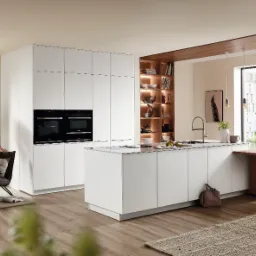 Jasna, przestronna kuchnia z białymi, minimalistycznymi szafkami, wyspą kuchenną i drewnianymi akcentami. W tle kobieta siedząca w fotelu, relaksująca się z napojem. Widok z okna na budynek...