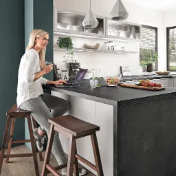 Kobieta siedzi na drewnianym stołku przy czarnym blacie w kuchni w Katowicach, korzystając z laptopa i trzymając filiżankę, z owocami i dekoracjami w tle.