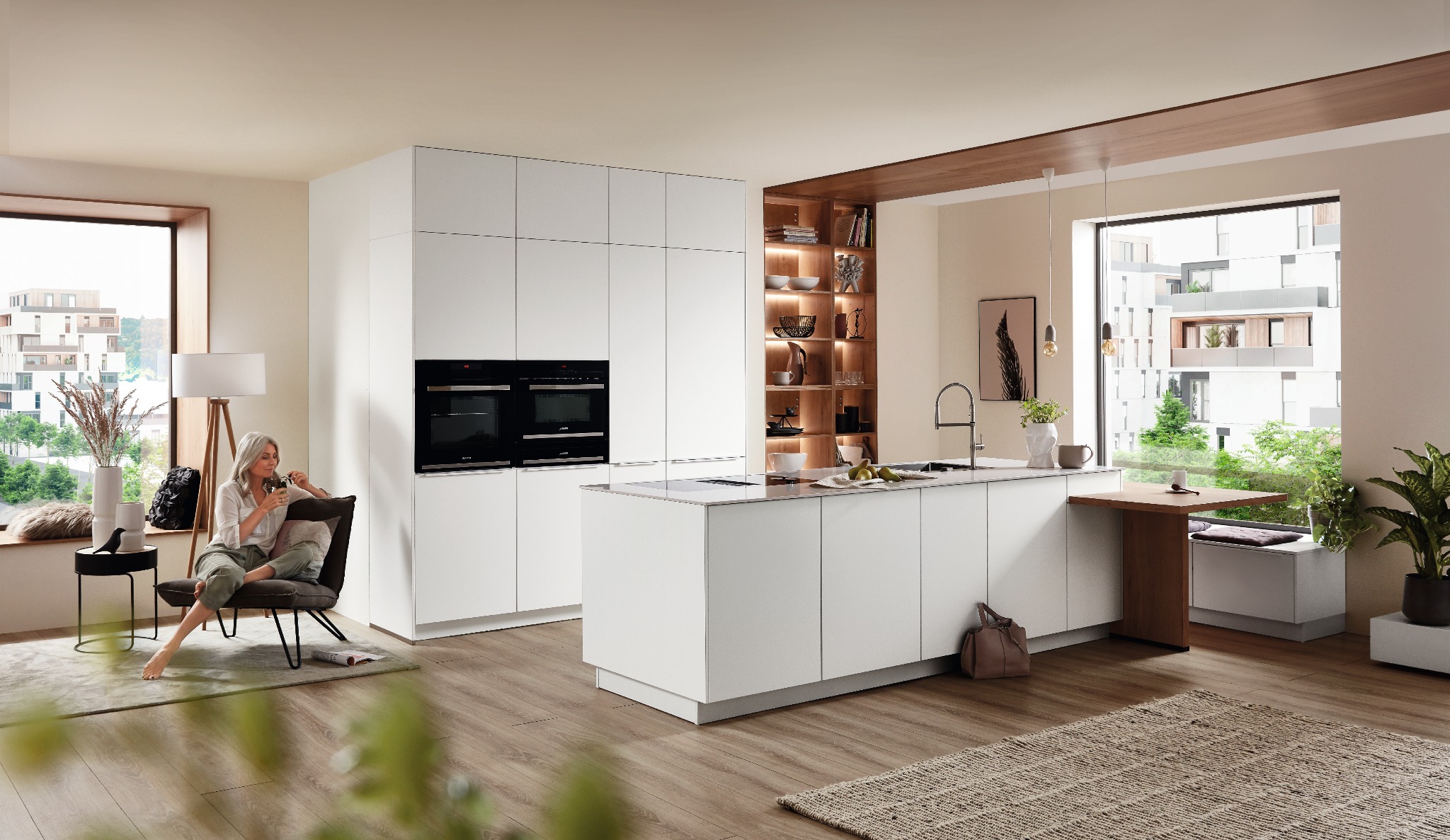 Jasna, przestronna kuchnia z białymi, minimalistycznymi szafkami, wyspą kuchenną i drewnianymi akcentami. W tle kobieta siedząca w fotelu, relaksująca się z napojem. Widok z okna na budynek...