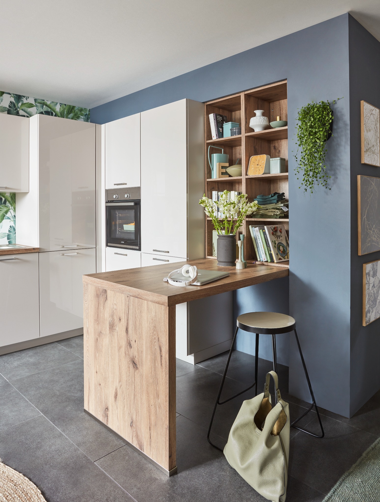 Nowoczesna kuchnia z białymi szafkami, drewnianym blatem roboczym pełniącym funkcję barku, otwartą półką z dekoracjami i niebieską ścianą. Na blacie laptop i słuchawki.