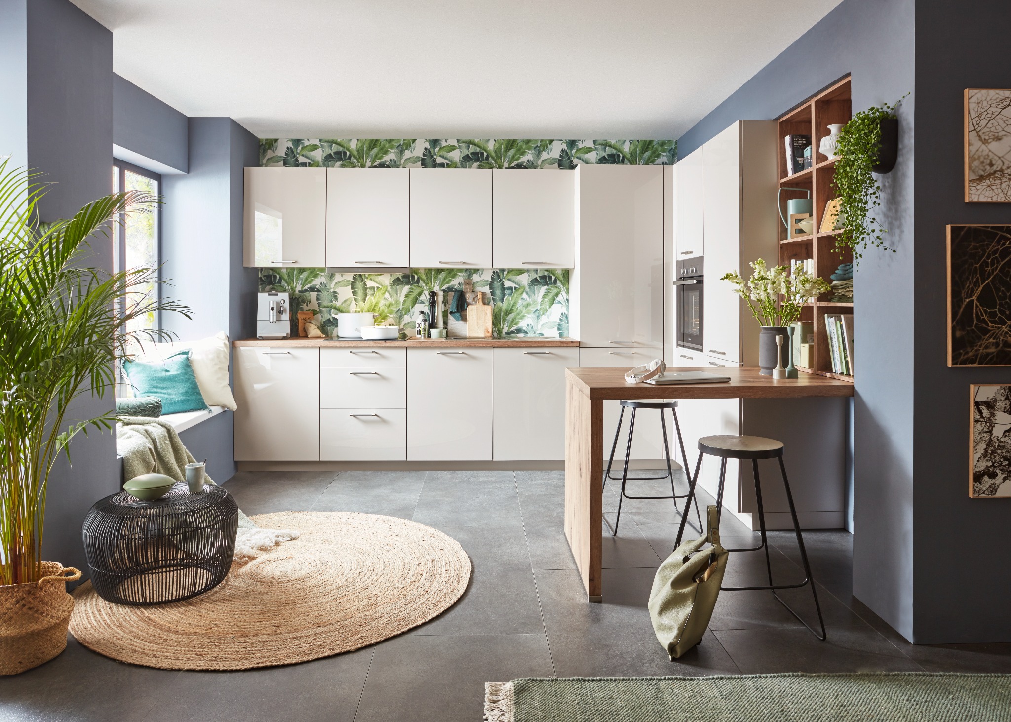 Jasna kuchnia z białymi szafkami i blatem z jasnego drewna, wzorzysta tapeta z motywem liści, okrągły dywan z juty i wysokie krzesła barowe, uzupełniona roślinami doniczkowymi i elementami...