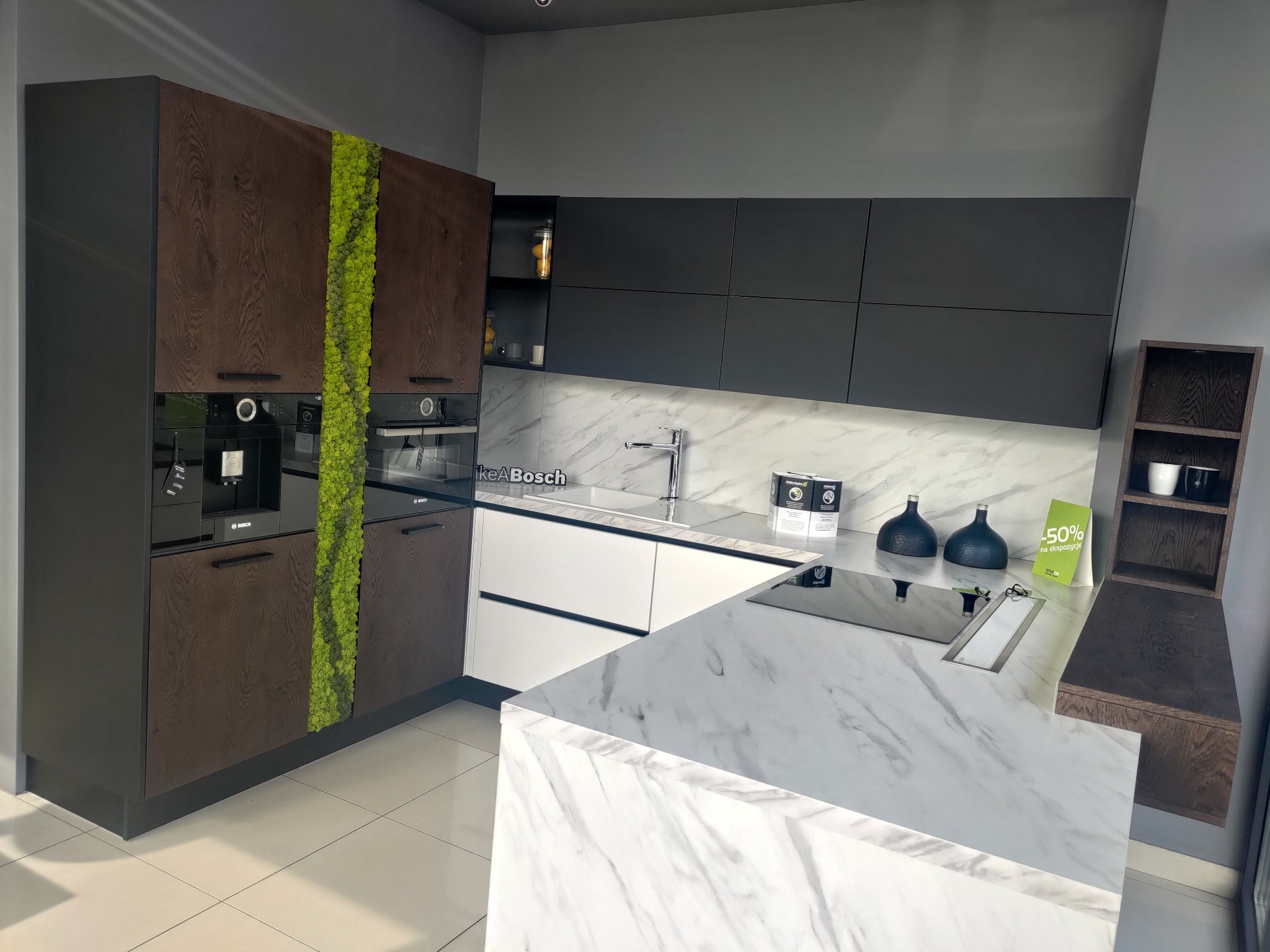 Nowoczesna kuchnia z ciemnymi, drewnopodobnymi szafkami, białymi frontami dolnymi i marmurowym blatem, z pionowym pasem mchu dekoracyjnego na szafce i wbudowanymi urządzeniami marki Bosch.