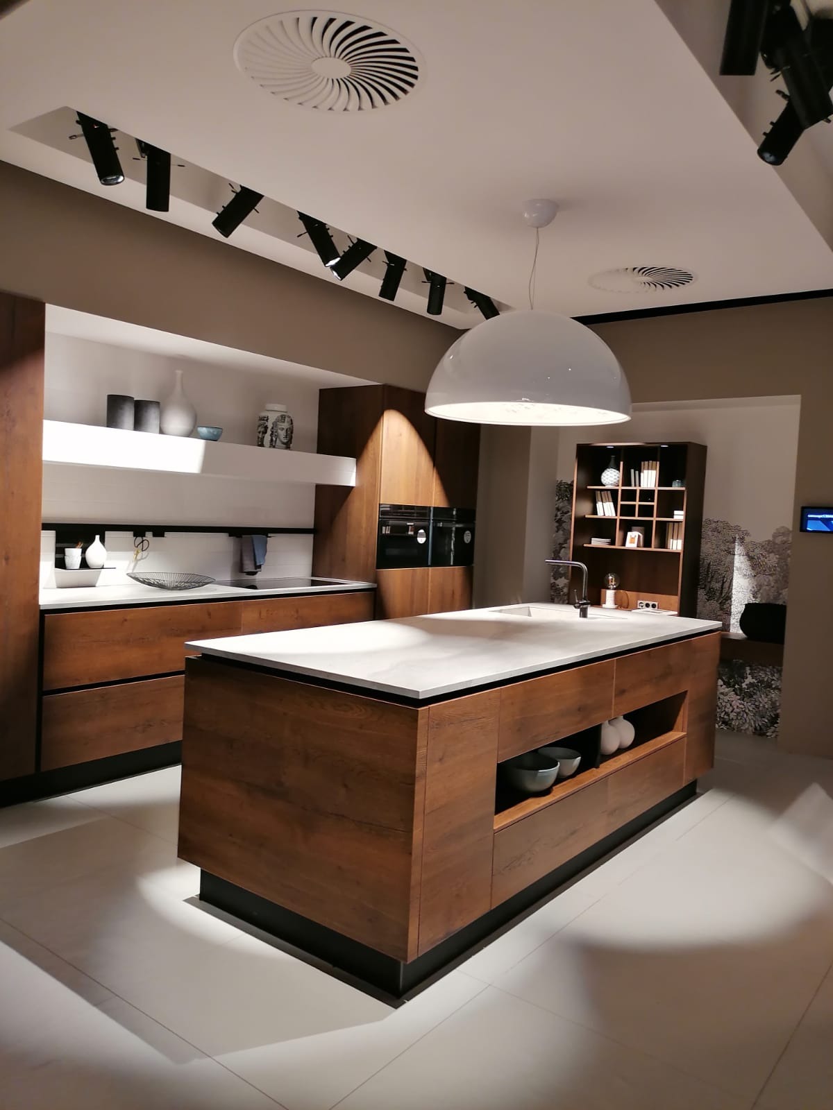 Nowoczesna kuchnia z wyspą, drewniane fronty szafek z wyraźnym usłojeniem, biały blat wyspy, okrągła lampa sufitowa, oświetlenie punktowe na szynie, dekoracyjna tapeta w tle.