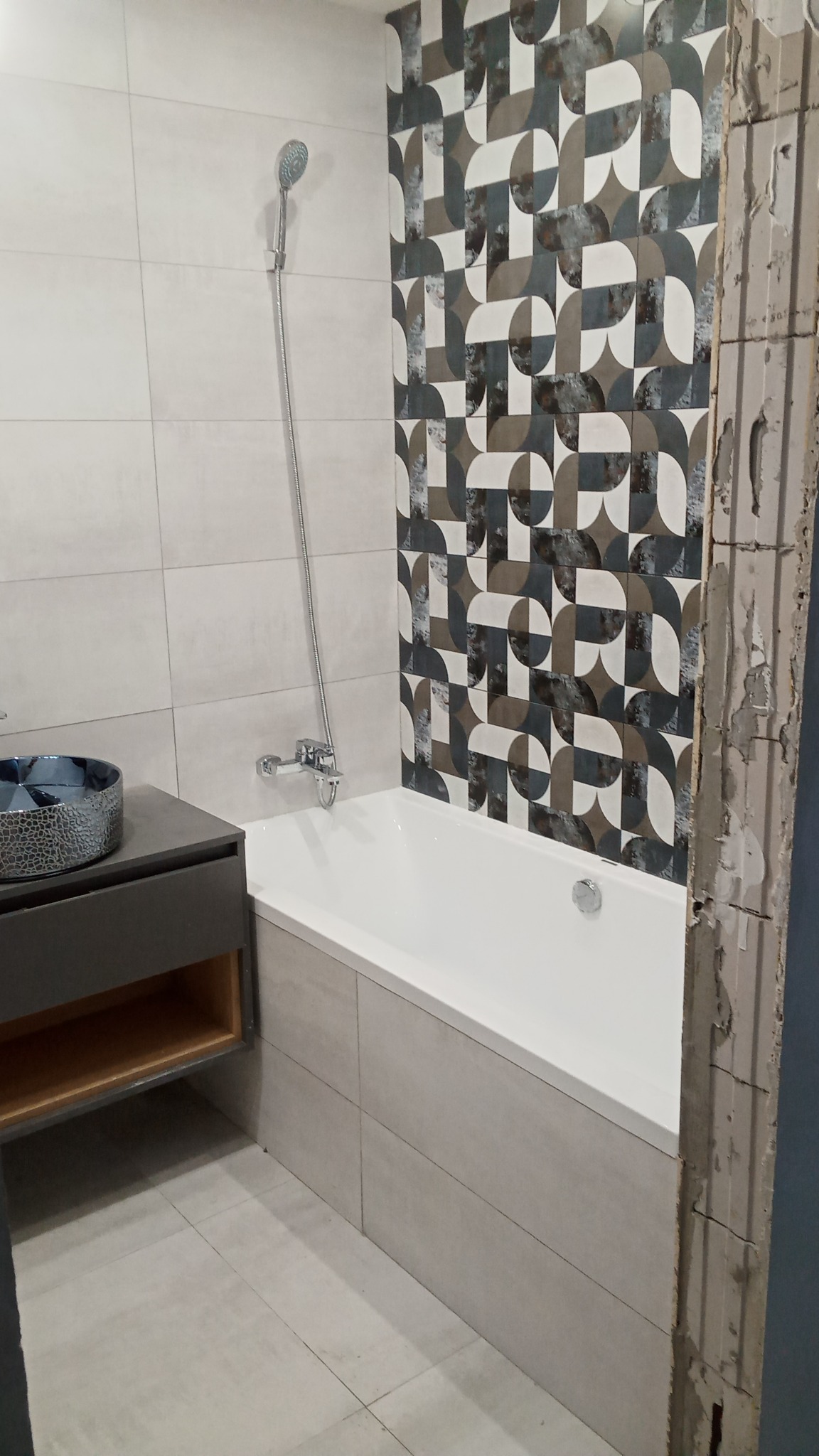 Nowoczesna łazienka z wanną obudowaną płytkami w odcieniach szarości, umywalką nablatową w metalicznym odcieniu oraz ścianą z geometrycznym wzorem płytek, widoczny fragment ściany w stanie surowym.