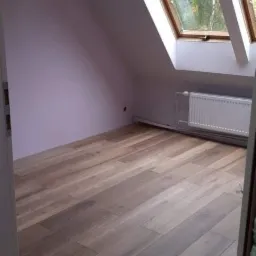 Poddasze z drewnianą podłogą, skośnym oknem dachowym i świeżo pomalowaną ścianą w odcieniach lilii, widoczny grzejnik pod oknem.