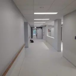 Długi, pomalowany na szaro korytarz z białymi panelami sufitowymi i drewnianą poręczą, widziany z perspektywy końca korytarza.
