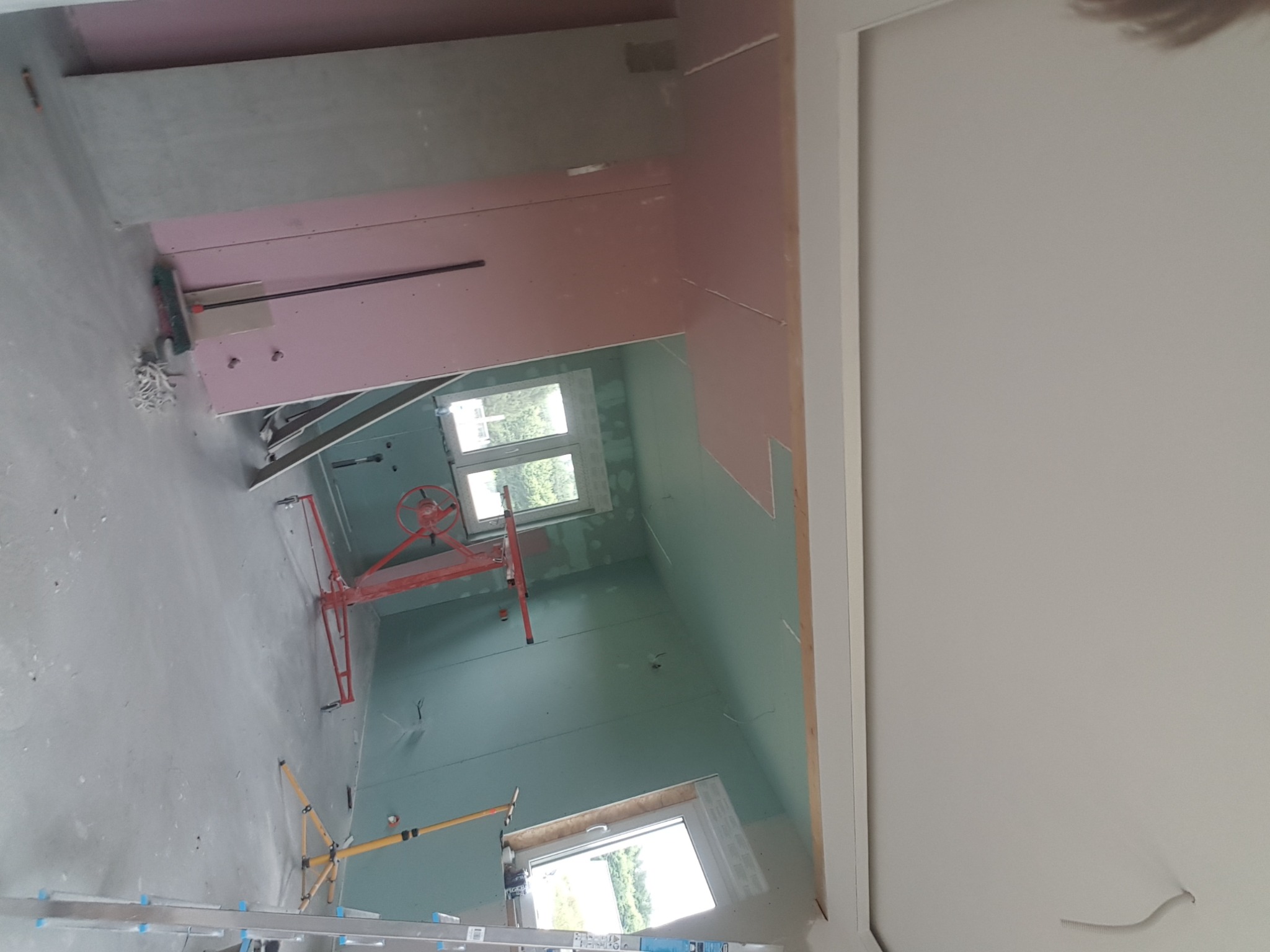 Wnętrze w trakcie montażu płyt kartonowo-gipsowych na poddaszu, widoczne stelaże, podnośnik do płyt, okna dachowe i częściowo zamontowane płyty w kolorze różowym i zielonym.