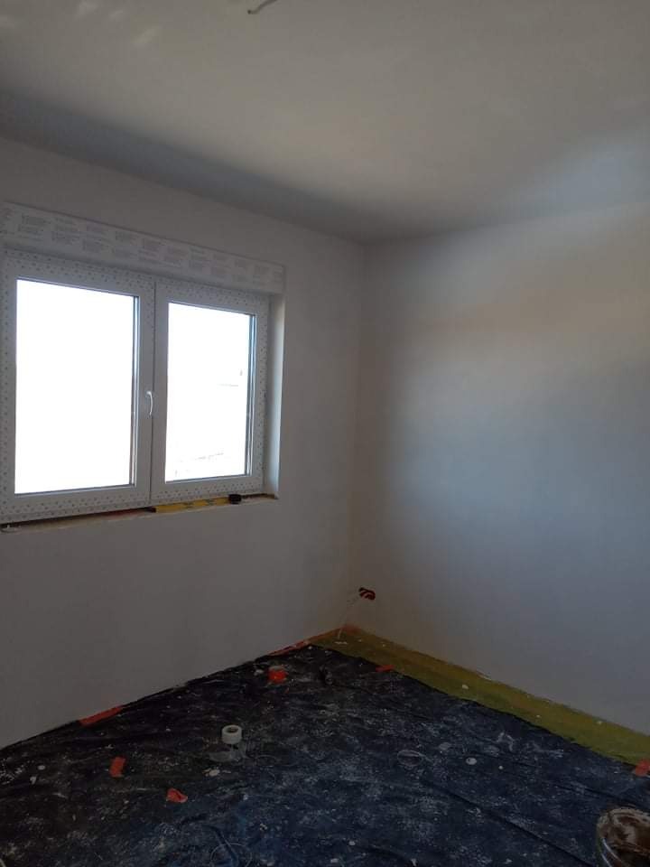 Pokój po gładziowaniu ścian, przygotowany do malowania, z zabezpieczoną podłogą folią malarską. Widoczne gniazdko elektryczne i okno z roletą.