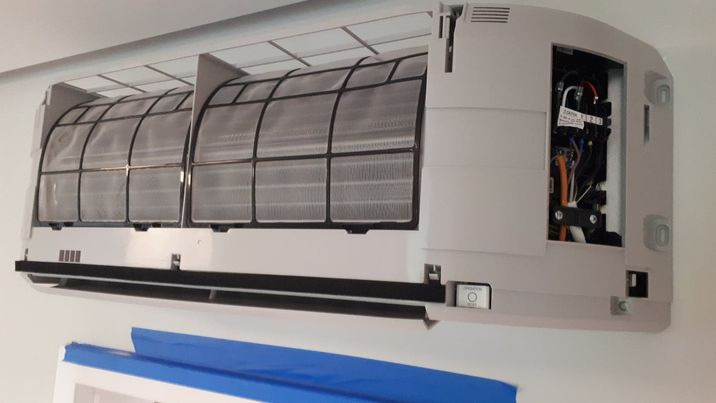 Otwarty panel klimatyzatora ściennego z widocznymi filtrami i wewnętrzną elektroniką, dolna krawędź maskowana niebieską taśmą malarską.