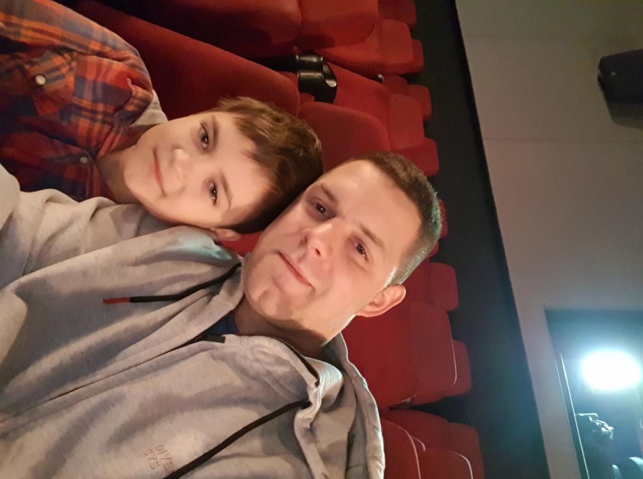 Mężczyzna z synem w kinie, siedzą na czerwonych fotelach. Uśmiechnięci, w swobodnych ubraniach. Widoczny fragment ekranu i oświetlenie.