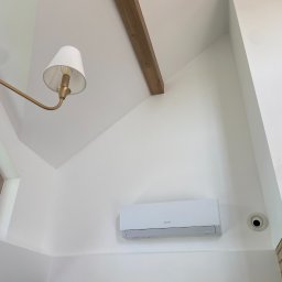 KLIMATIK-VENT MATEUSZ CISZ - Biała klimatyzacja split marki Gree zamontowana na białej ścianie pod skosem, widok od dołu z fragmentem ozdobnej lampy i drewnianej belki stropowej.