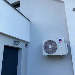 Zewnętrzna jednostka klimatyzacji marki Gree zamontowana na jasnoszarym elewacji budynku z widocznymi elementami architektonicznymi, takimi jak okno w kształcie trójkąta i reflektor nad drzwiami.