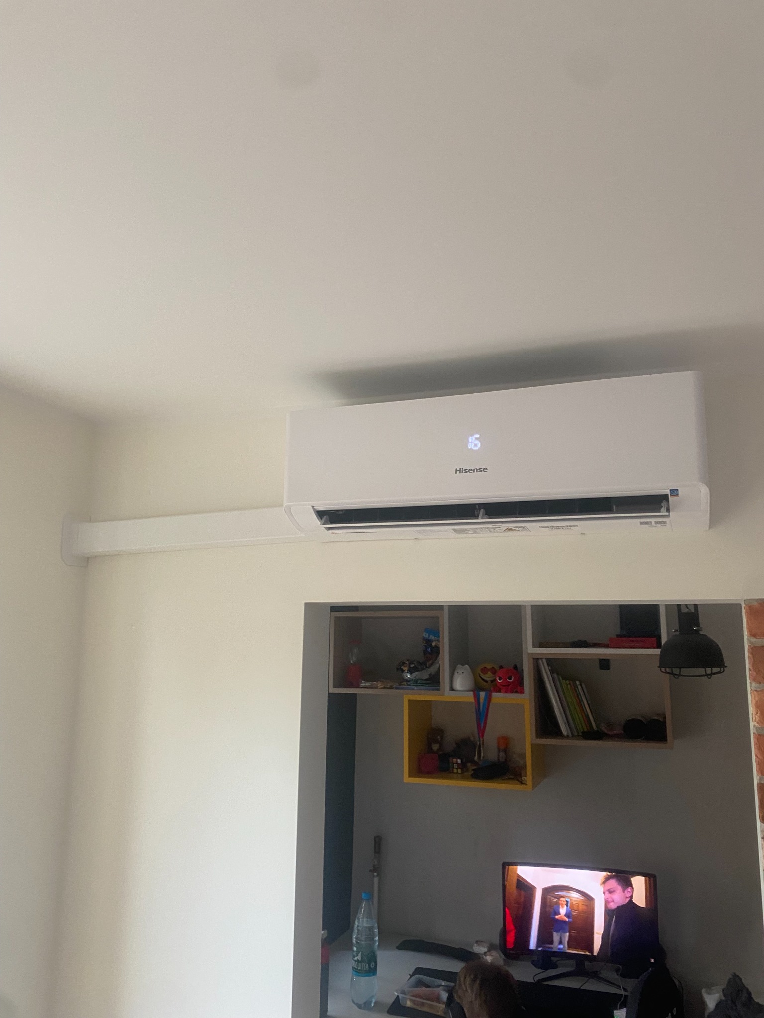 Biały klimatyzator Hisense zamontowany na ścianie w pokoju, wyświetlający temperaturę 16 stopni Celsjusza, z widoczną półką z dekoracjami i włączonym telewizorem w tle.