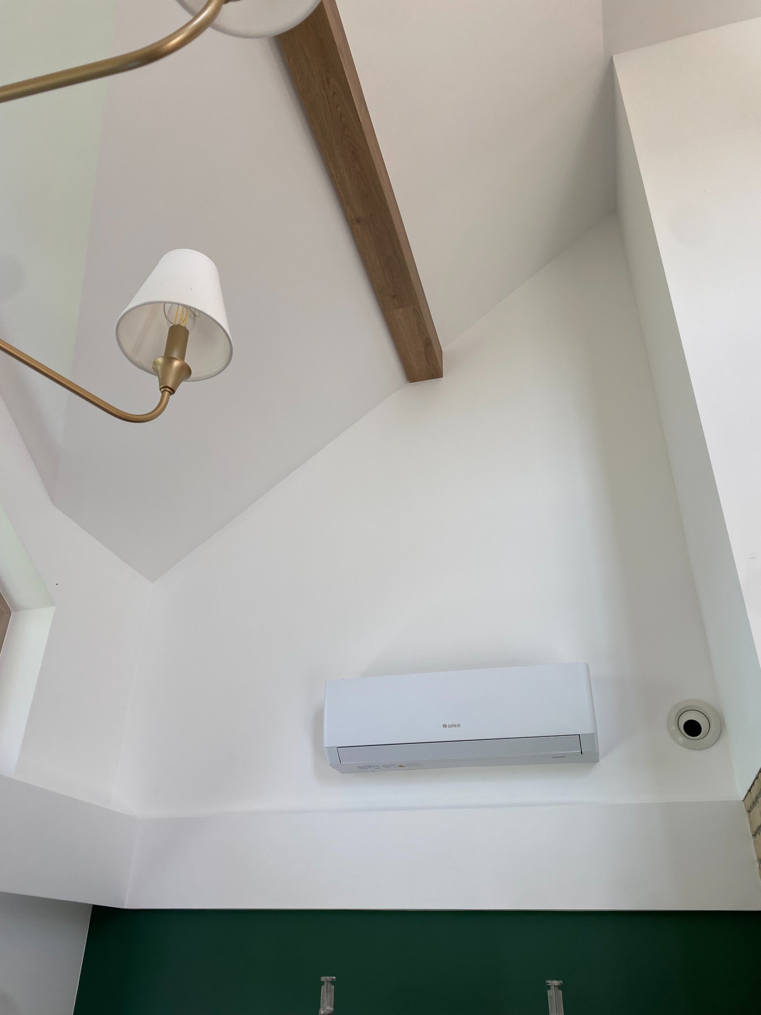 Biała klimatyzacja split marki Gree zamontowana na białej ścianie pod skosem, widok od dołu z fragmentem ozdobnej lampy i drewnianej belki stropowej.