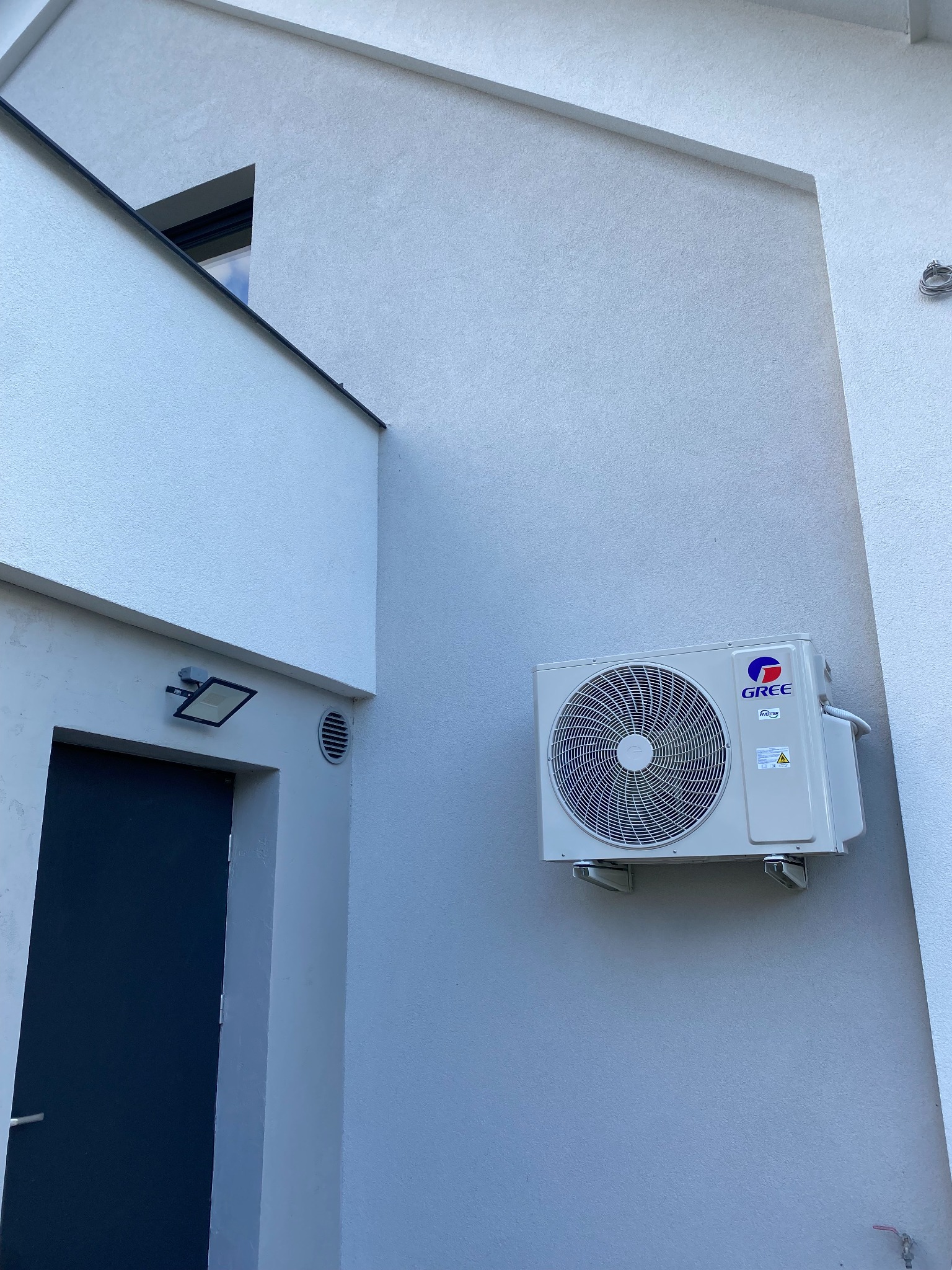 Zewnętrzna jednostka klimatyzacji marki Gree zamontowana na jasnoszarym elewacji budynku z widocznymi elementami architektonicznymi, takimi jak okno w kształcie trójkąta i reflektor nad drzwiami.